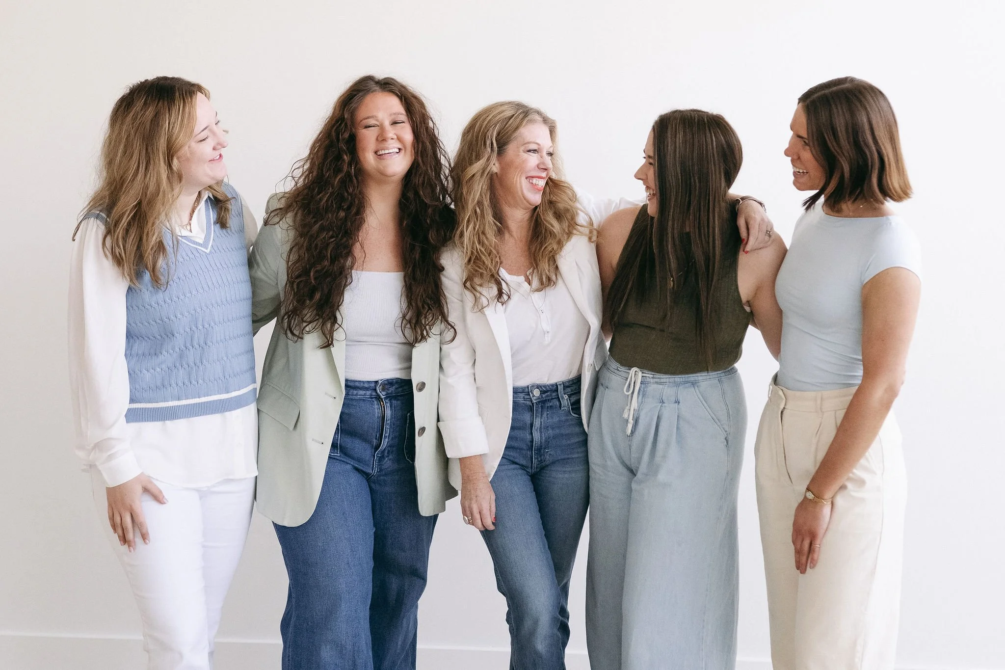 Five women standing together, smiling and laughing in front of a plain white wall, dressed casually.