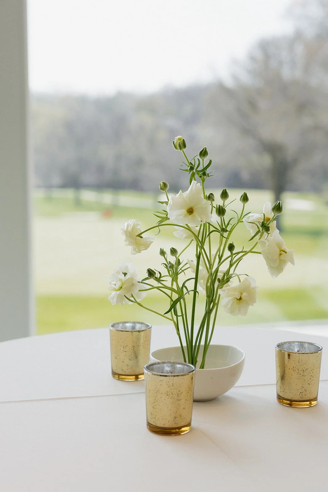 A white table with a white bowl of white flowers and three small candles in gold holders, set against a window showing an outdoor landscape of grass and trees.