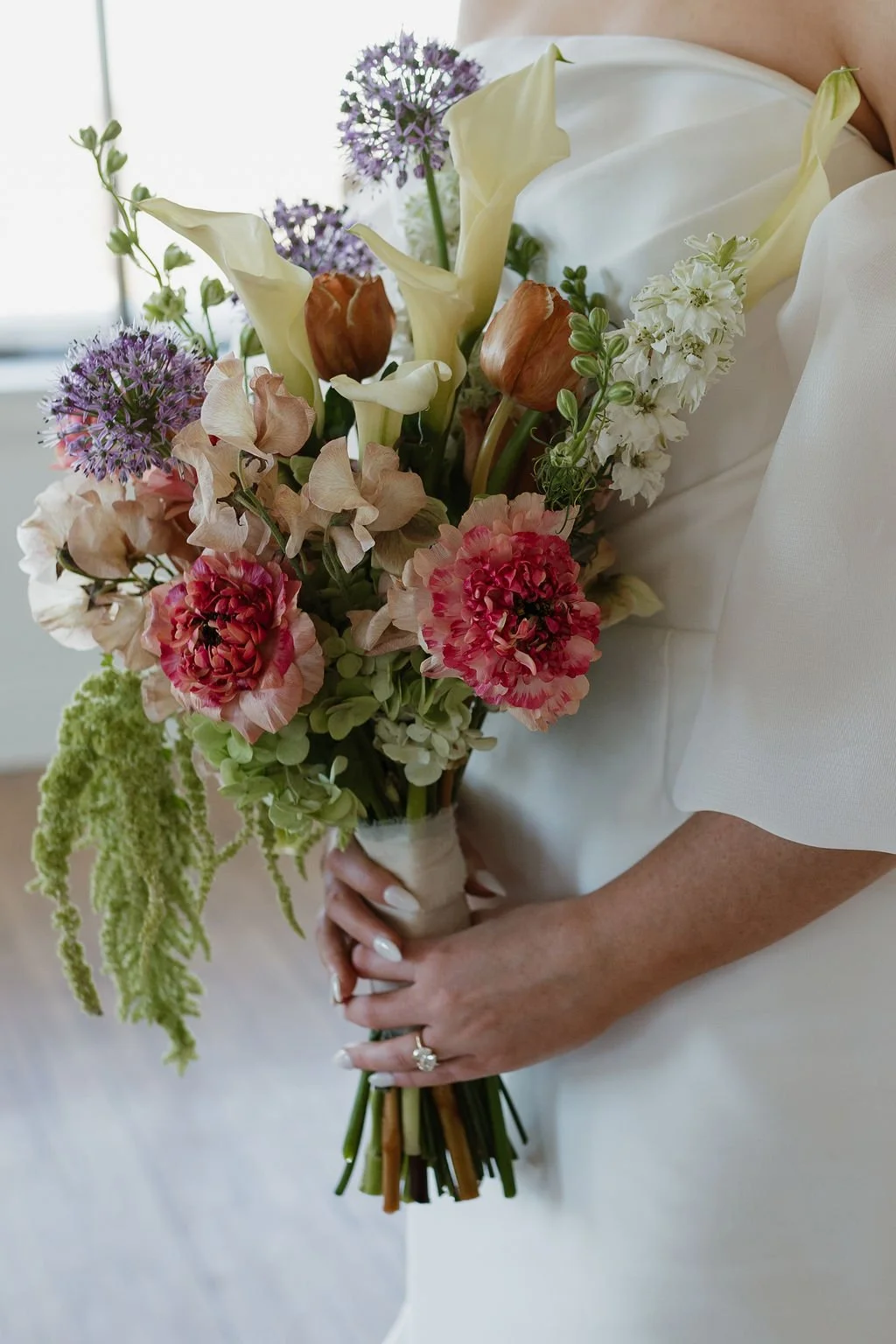Person holding a bouquet of mixed flowers, including calla lilies, hydrangeas, peonies, and tulips, against a blurred indoor background.
