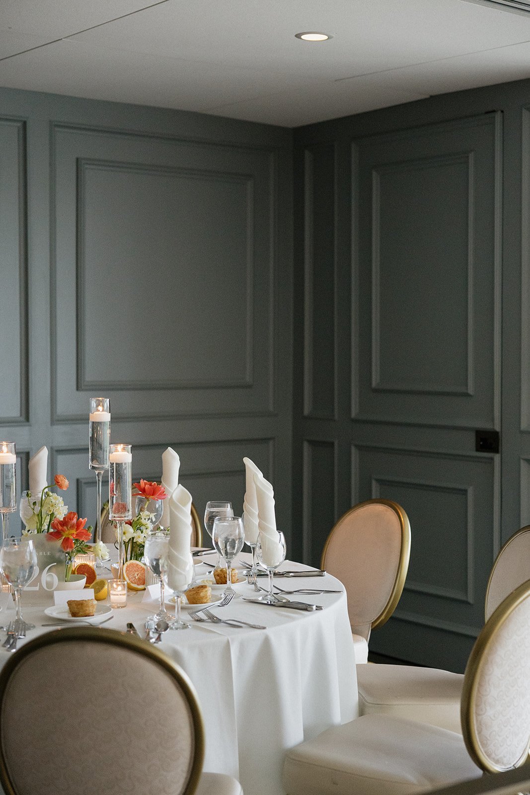 Elegant dining table set for a formal event with white tablecloth, folded napkins, glassware, flowers, candles, and small appetizers in a room with gray paneled walls.