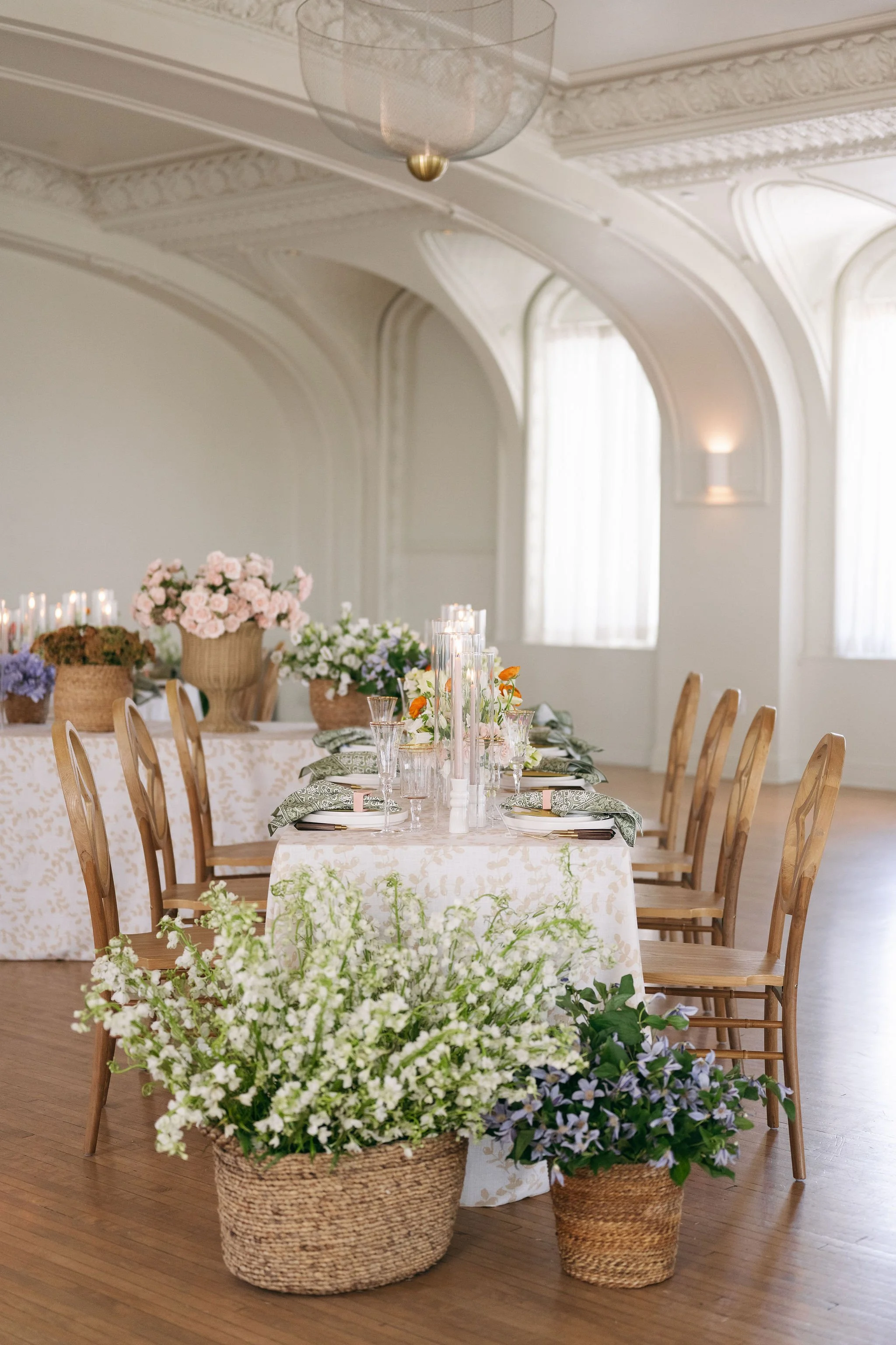 Elegant dining table set for a special occasion with floral centerpieces and candles, surrounded by wooden chairs on a polished wooden floor in a beautifully decorated room.