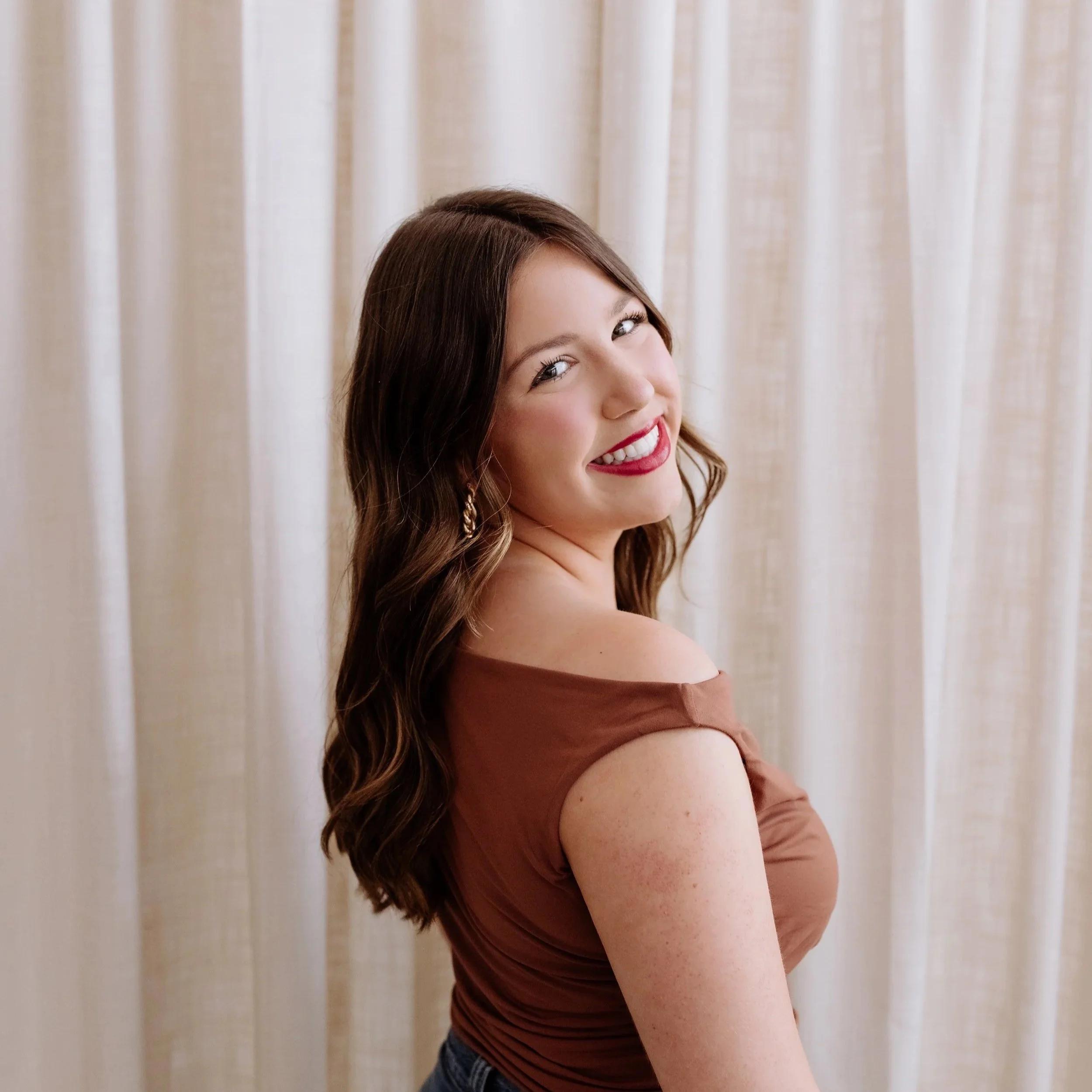 A young woman with short brown hair, blue eyes, and a nose piercing, smiling at the camera, wearing a white t-shirt, against a plain white background.