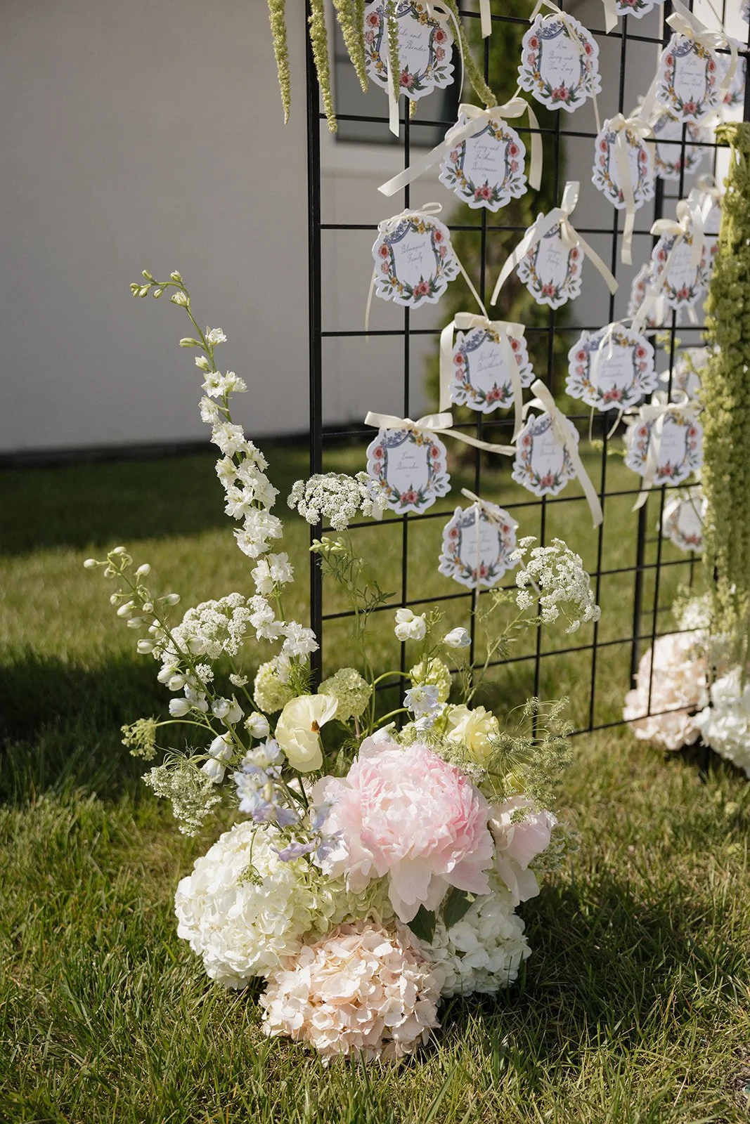 Decorative wedding or event seating chart with floral arrangement, featuring white, pink, and cream-colored flowers near a black grid display with floral and ribbon tags.