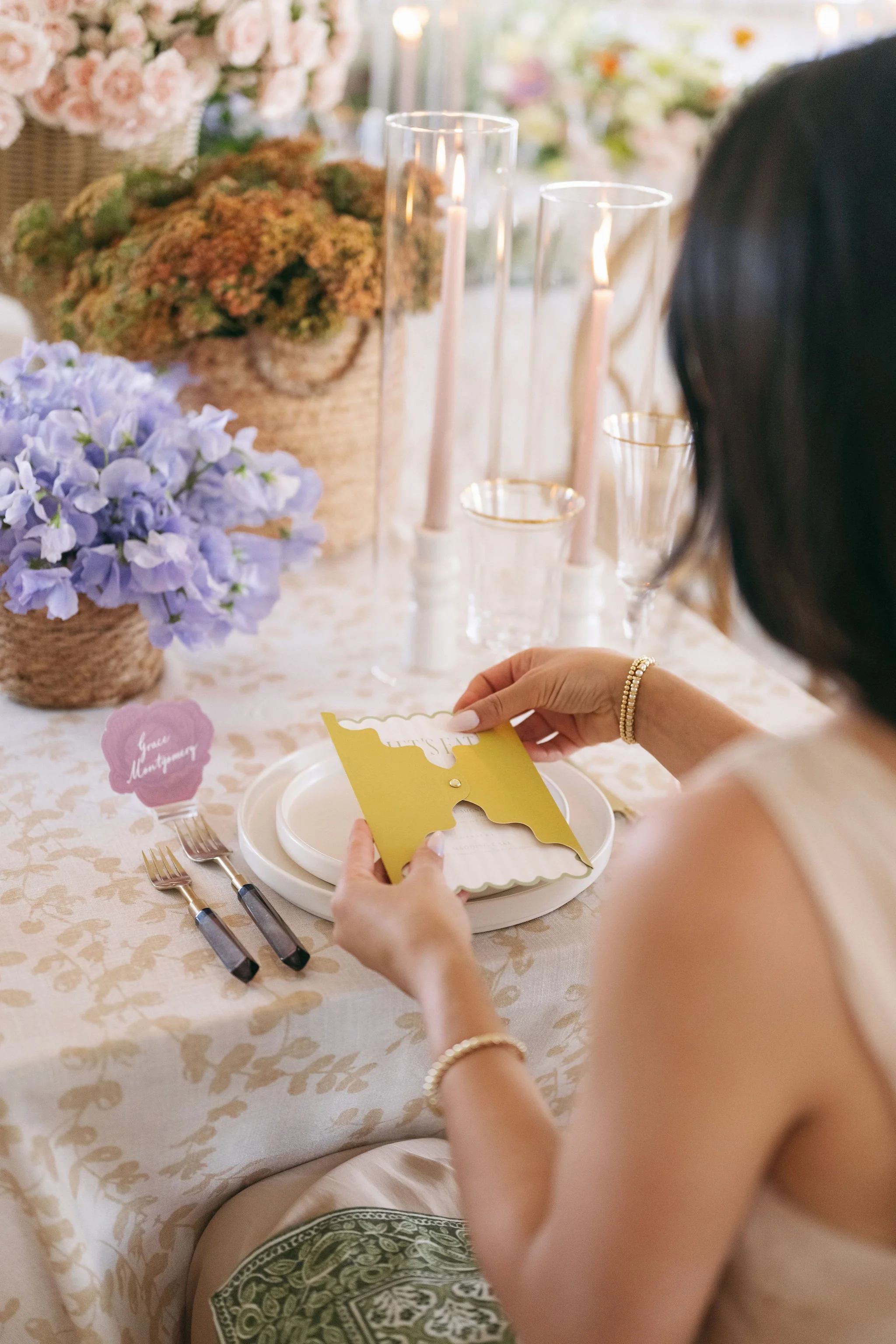 A woman at a decorated table is reading a yellow and white card. The table has floral arrangements, candles, and glassware. There is a small pink name tag reading 'Lisa Montgomery' next to her plate.