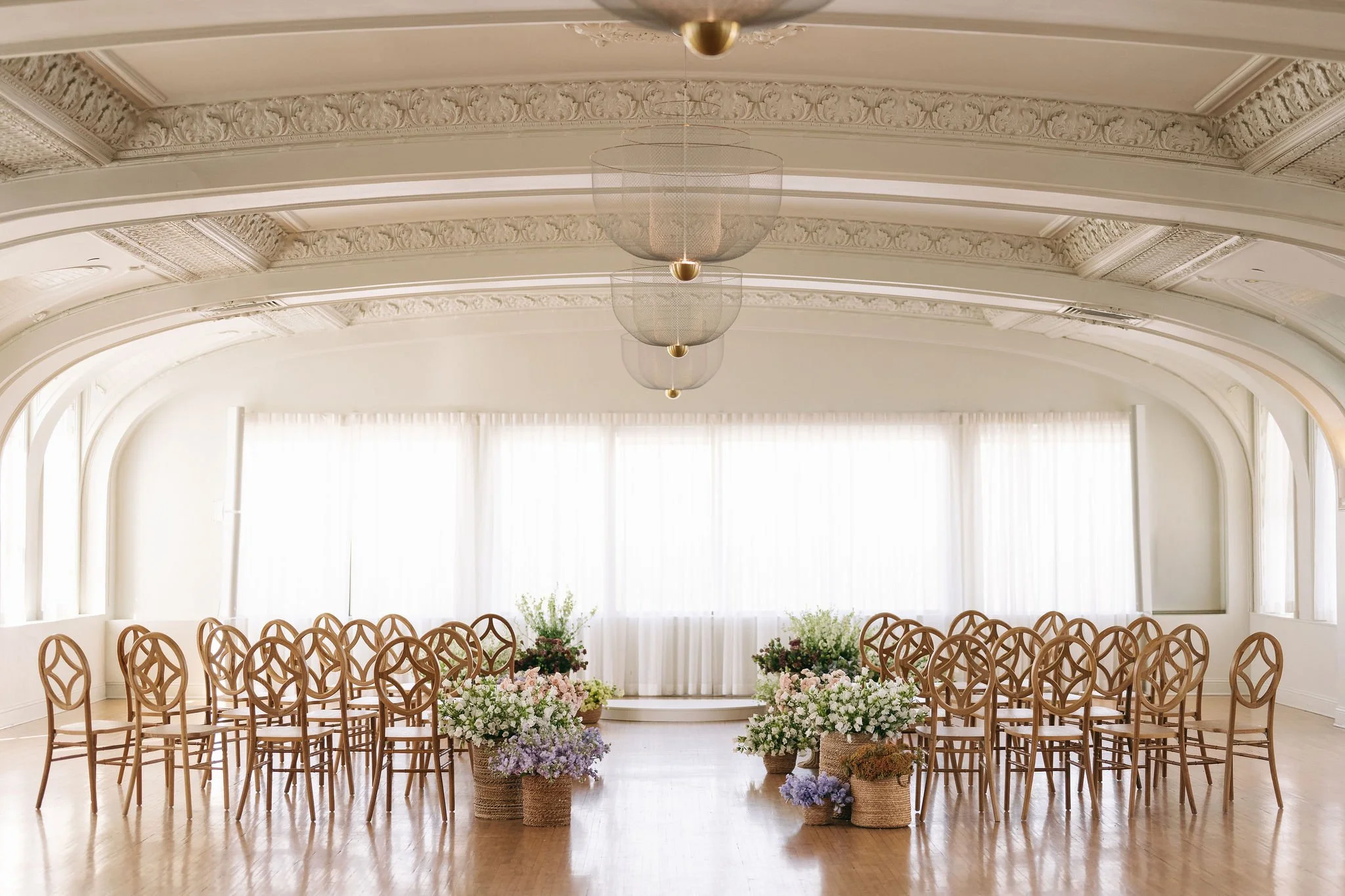 An elegant, white-walled room with ornate ceiling moldings and hanging lights, set up for a wedding or event with rows of wooden chairs and large floral arrangements in baskets.
