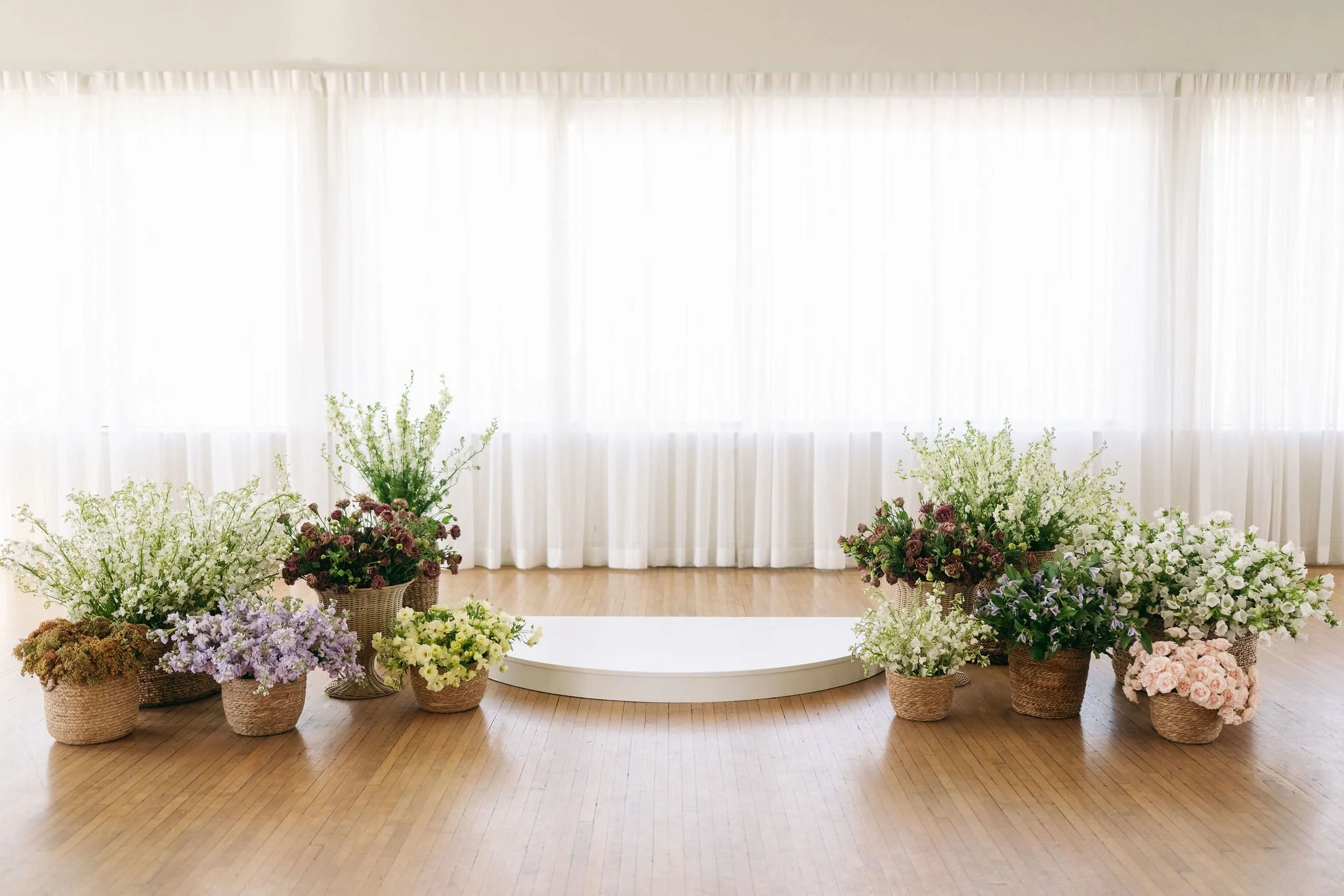 Empty stage with a white platform surrounded by flower arrangements in baskets and pots, in front of sheer white curtains
