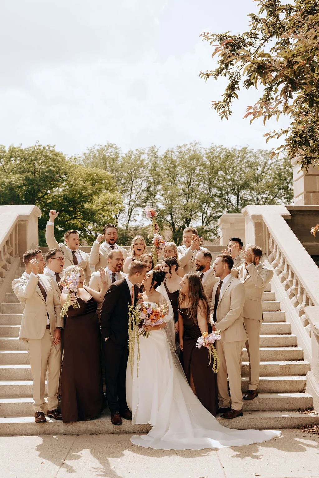 A wedding party on steps outdoors, with bride and groom at the center, surrounded by friends and family, celebrating with smiles and gestures on a sunny day.