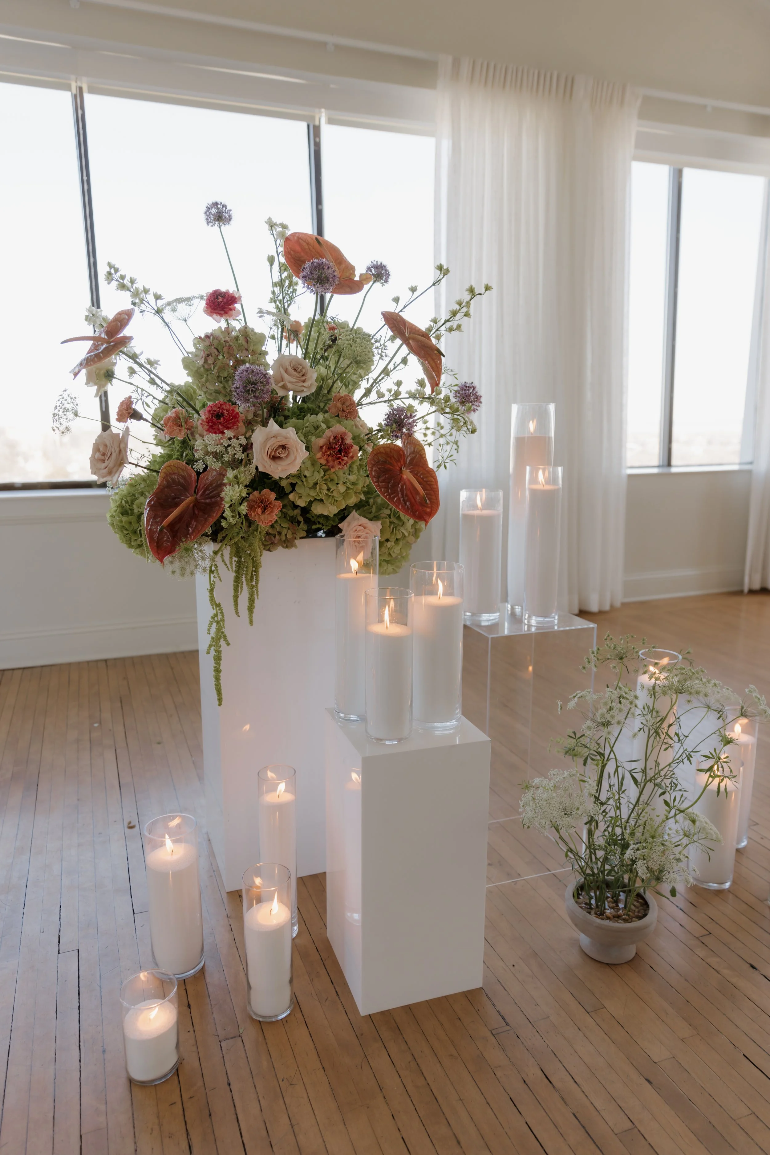 Elegant floral arrangement with pink and purple flowers and green foliage in a white vase, surrounded by tall candles in glass holders on a wooden floor with a large window and white curtains in the background.