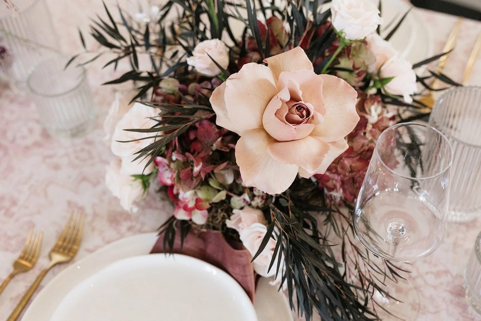 A floral centerpiece with cream and pink roses, purple and white flowers, and dark green leaves on a table set with a white plate, gold forks, and clear glassware.