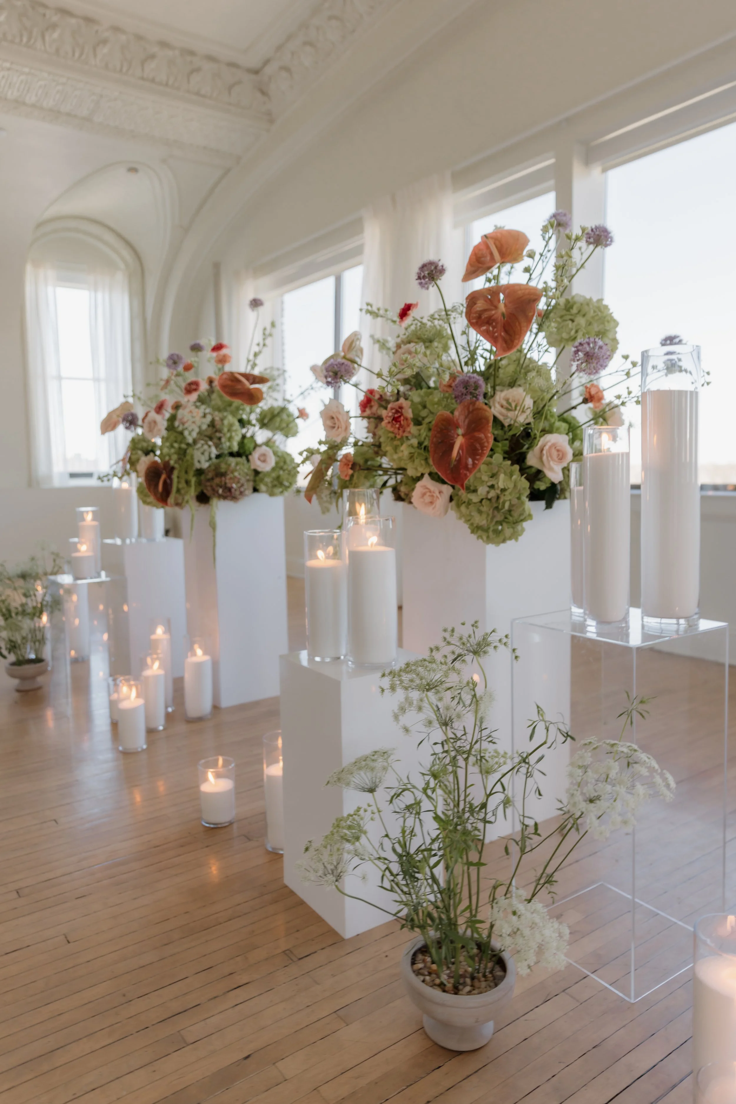 Elegant indoor floral display with large flower arrangements in white vases, surrounded by numerous lit candles on the wooden floor, in a bright room with decorative ceiling molding and large windows.