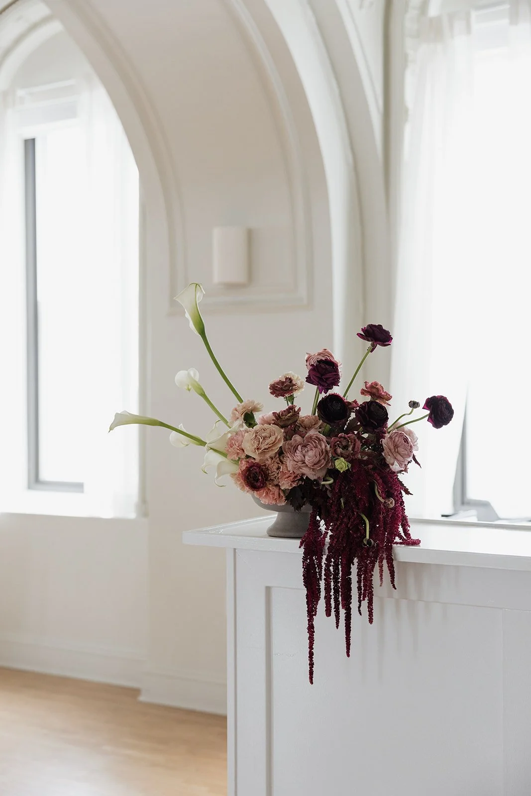 A floral arrangement with pink and dark purple flowers, including calla lilies and ranunculus, placed on a white shelf or cabinet in a bright room with white walls and windows.