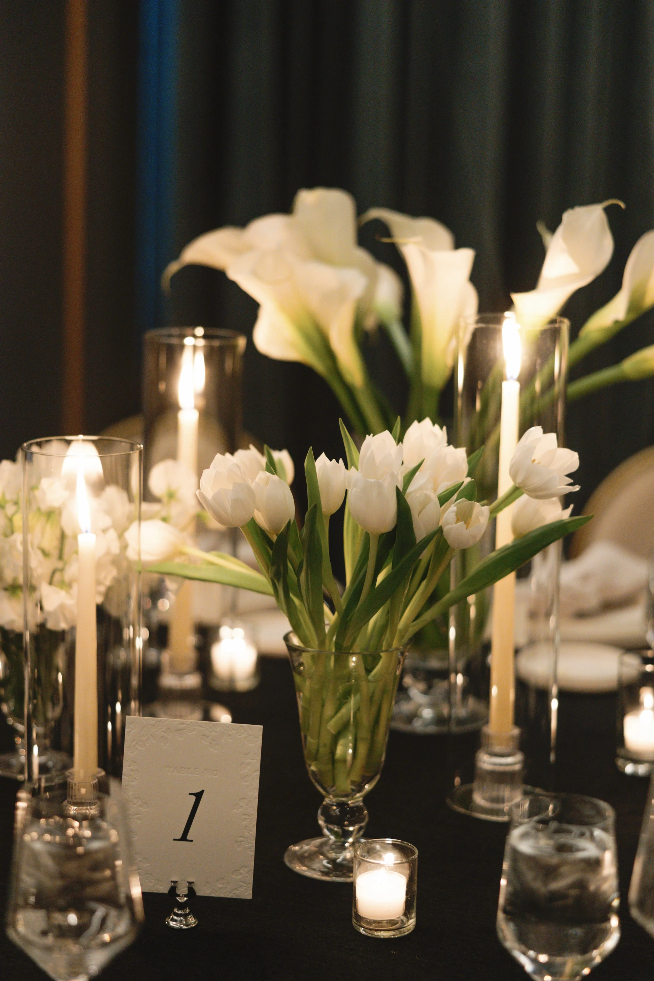 Elegant table setting with white lilies and tulips in a glass vase, lit candles in glass holders, and a table number card indicating Table 1.