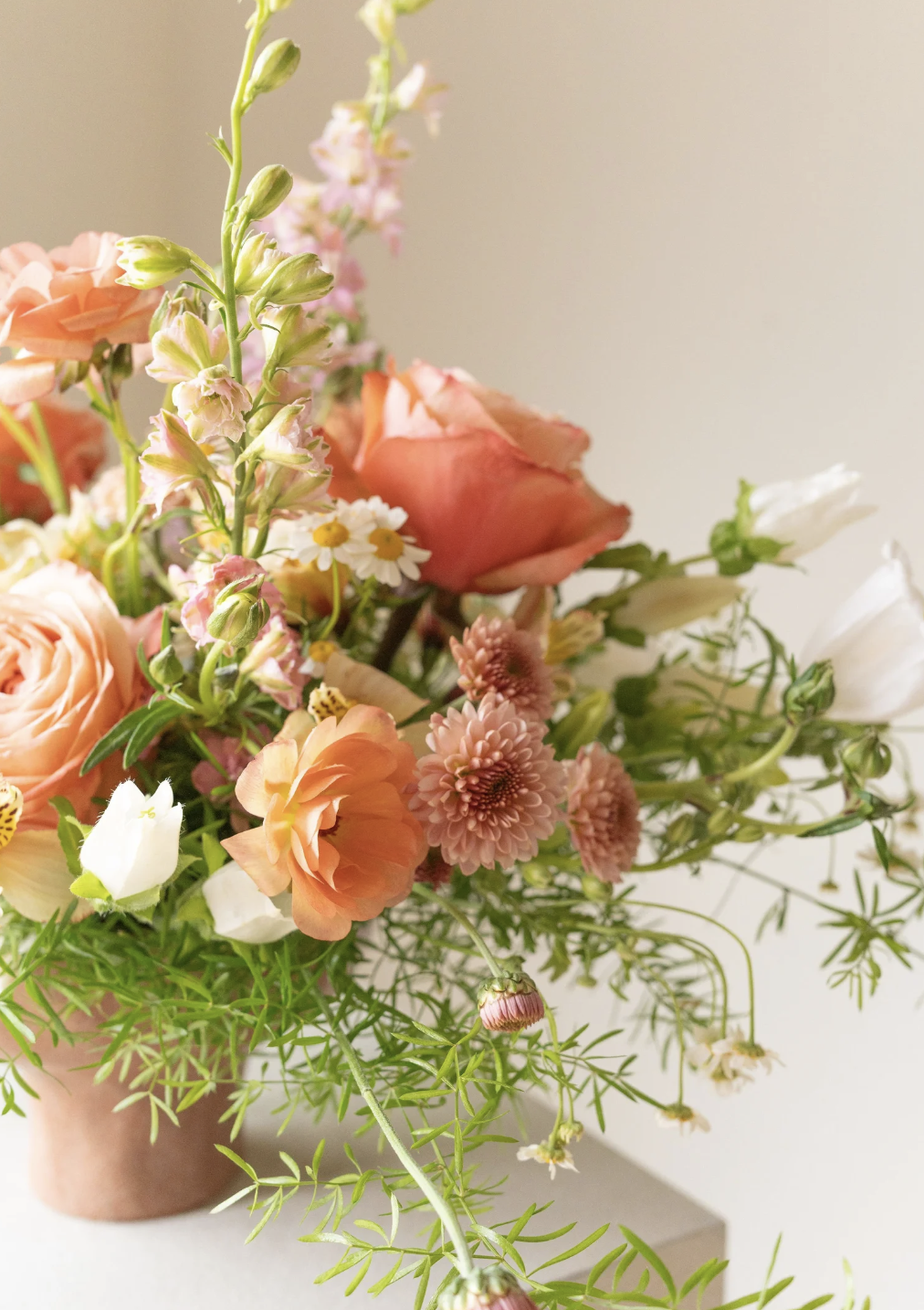 A bouquet of various flowers including pink roses, peach ranunculus, white daisies, and pink chrysanthemums in a terracotta pot.