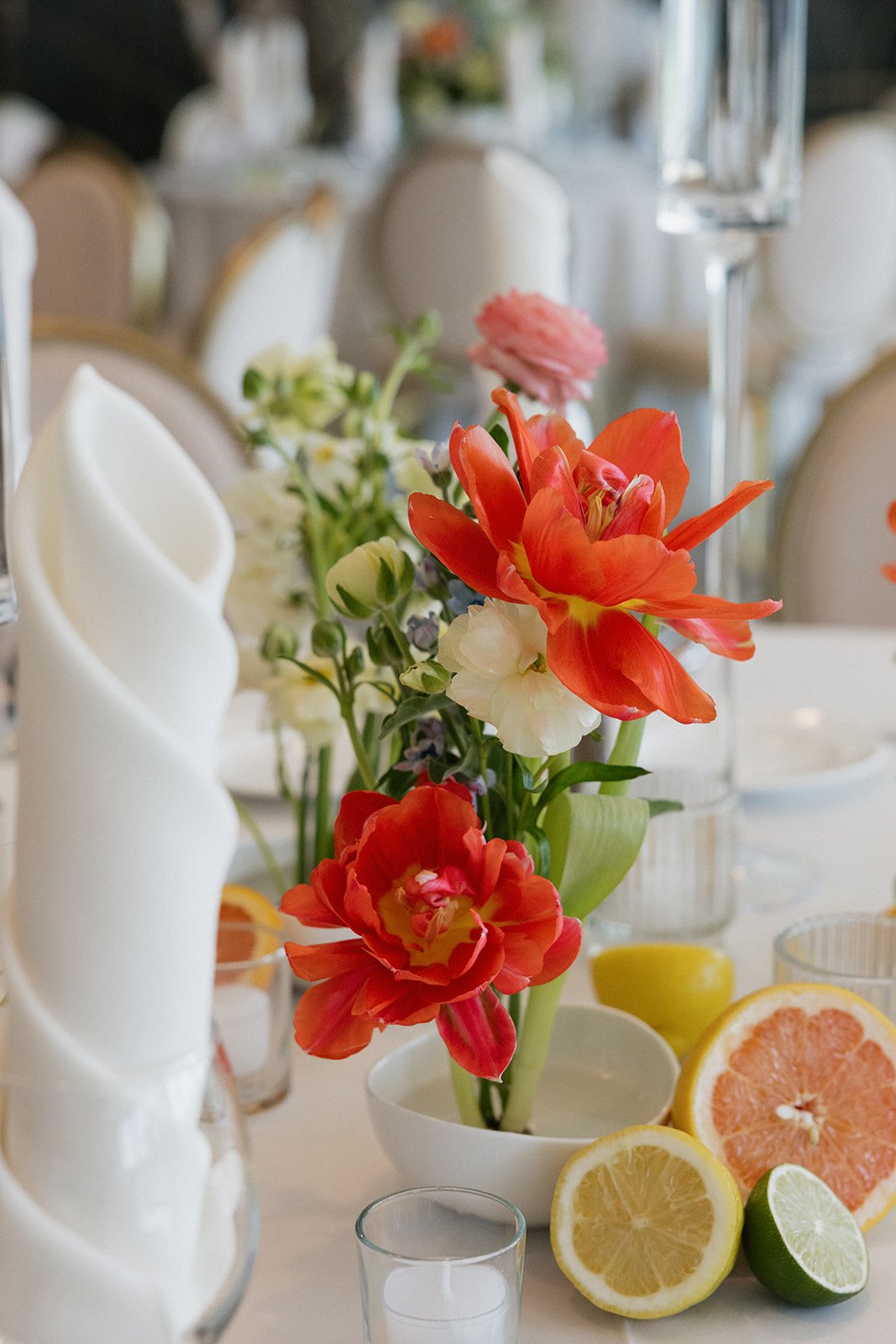 A floral centerpiece with red, white, and pink flowers on a table set for an event, with sliced lemons and grapefruit, glassware, and white napkins in the background.