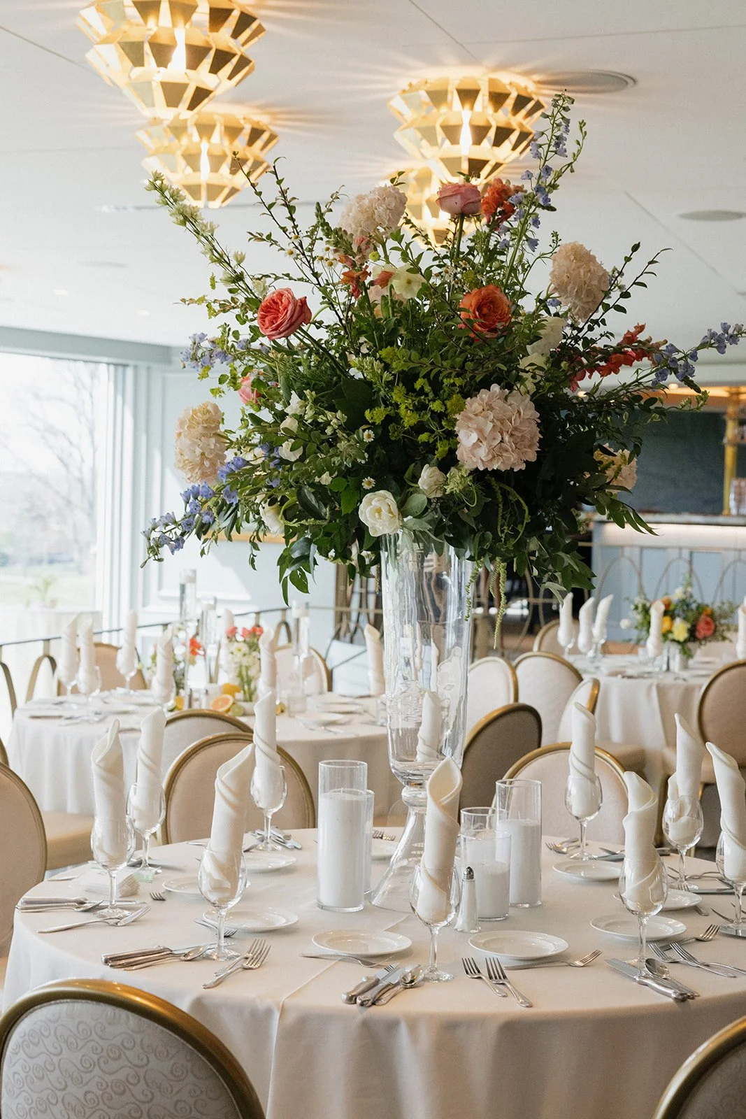 Elegant banquet table decorated with a large floral centerpiece in a glass vase, surrounded by neatly folded white napkins, candles, and silverware, set in a restaurant or event space with large windows and modern lighting