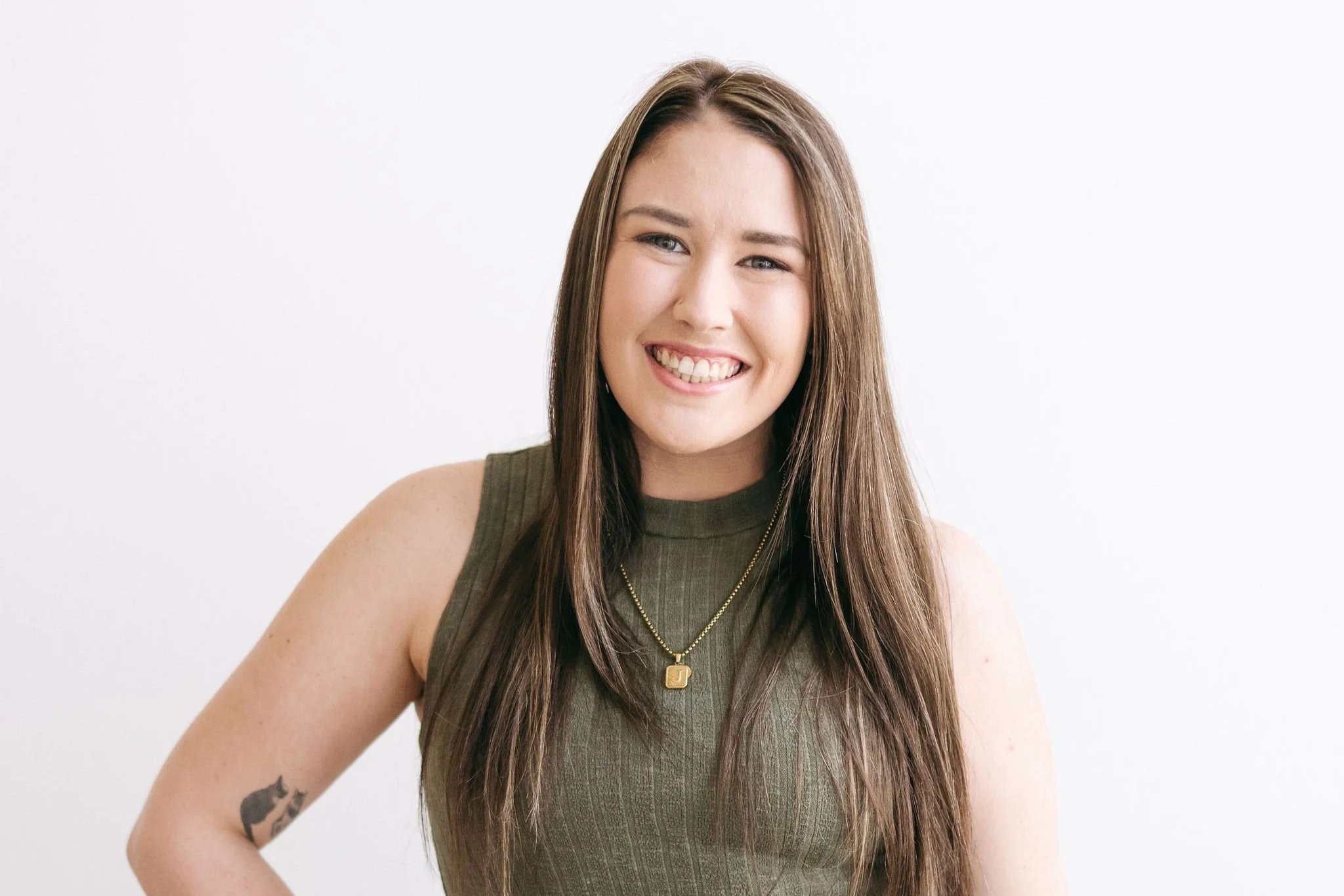 A young woman with long brown hair smiling at the camera, wearing a sleeveless green top, a gold necklace, and has a tattoo of a black cat on her left arm, standing against a plain white background.