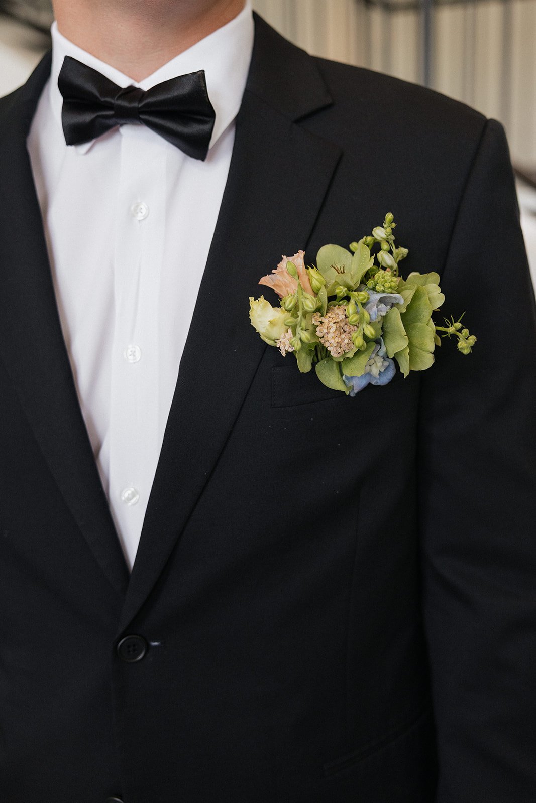 Close-up of a man wearing a black tuxedo with a white dress shirt and black bow tie, with a boutonniere made of pastel-colored flowers on his left lapel.