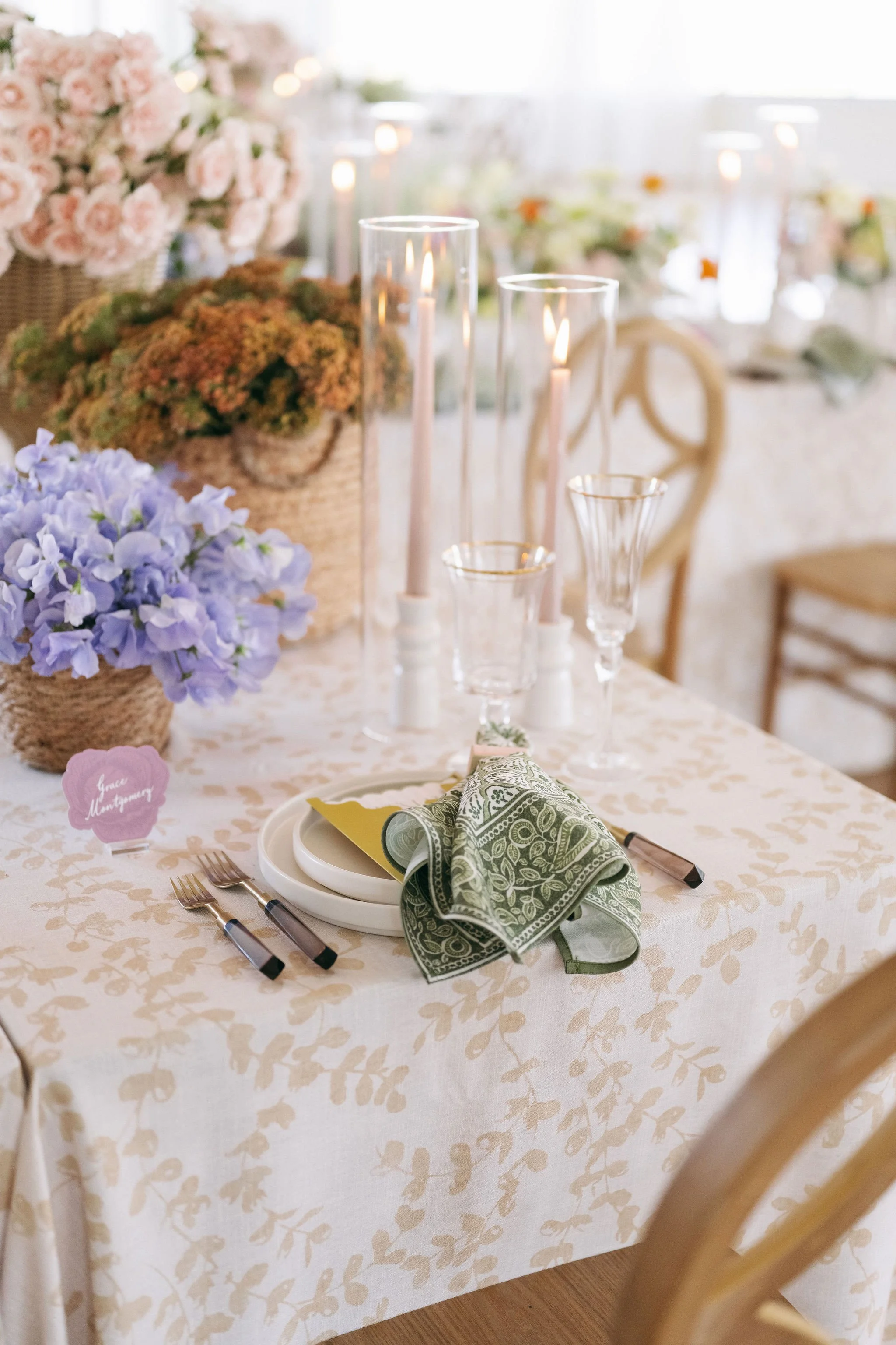 Elegant table setting for a wedding or special event featuring floral centerpieces, tall candles, glassware, plates, and napkins, with a handwritten name tag reading 'Fana Montgomery'.