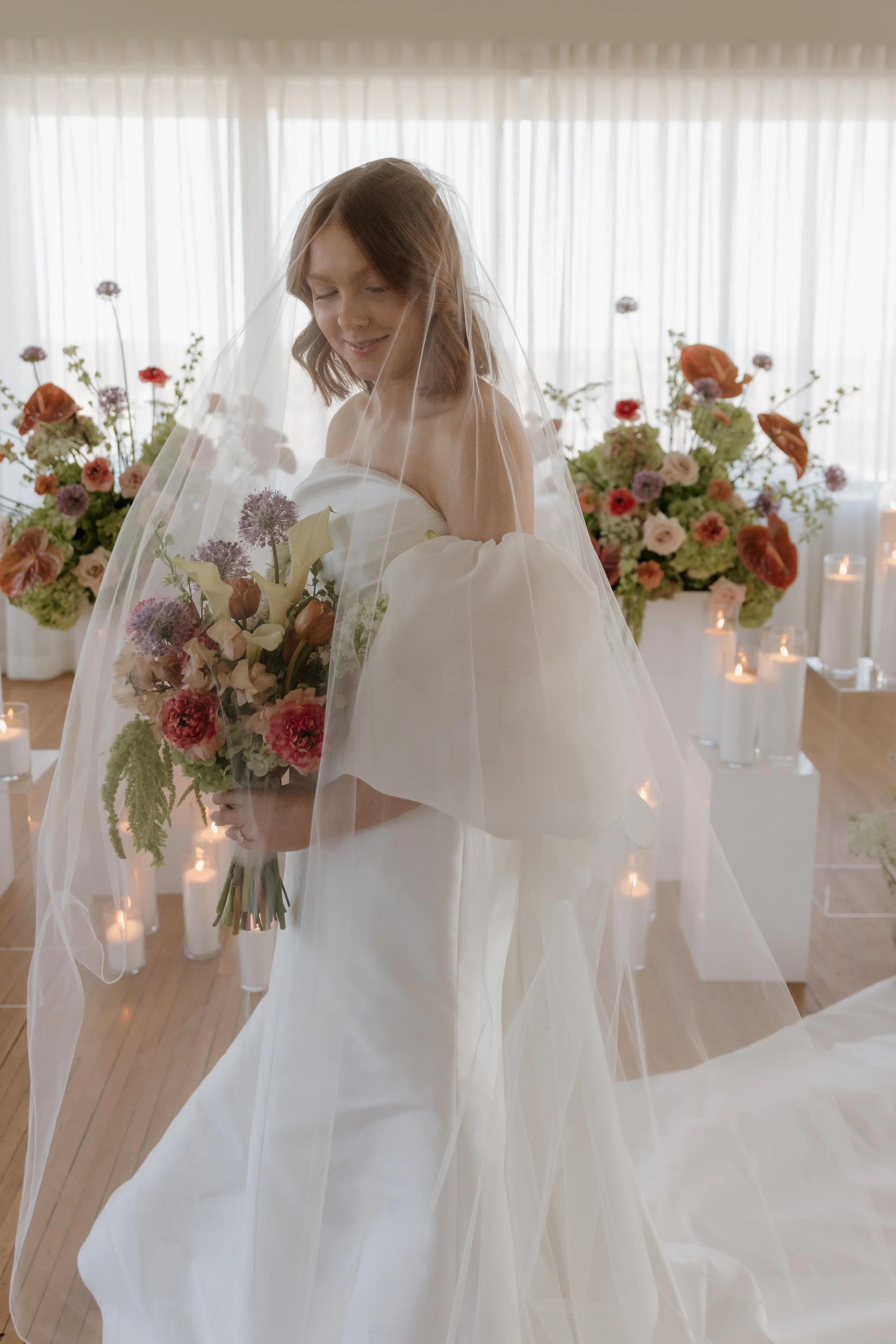 A bride with shoulder-length light brown hair smiling underneath a veil, holding a large bouquet of colorful flowers, surrounded by floral arrangements and candles.