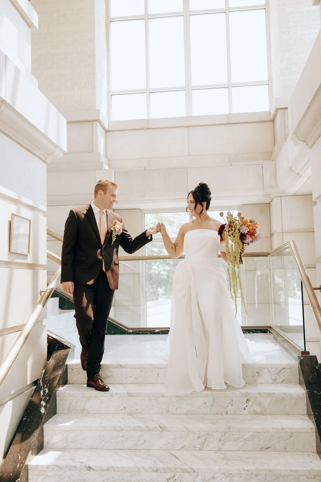 A bride and groom holding hands on a marble staircase in a bright, modern building with large windows, the bride in a white wedding dress holding a bouquet, and the groom in a dark suit.