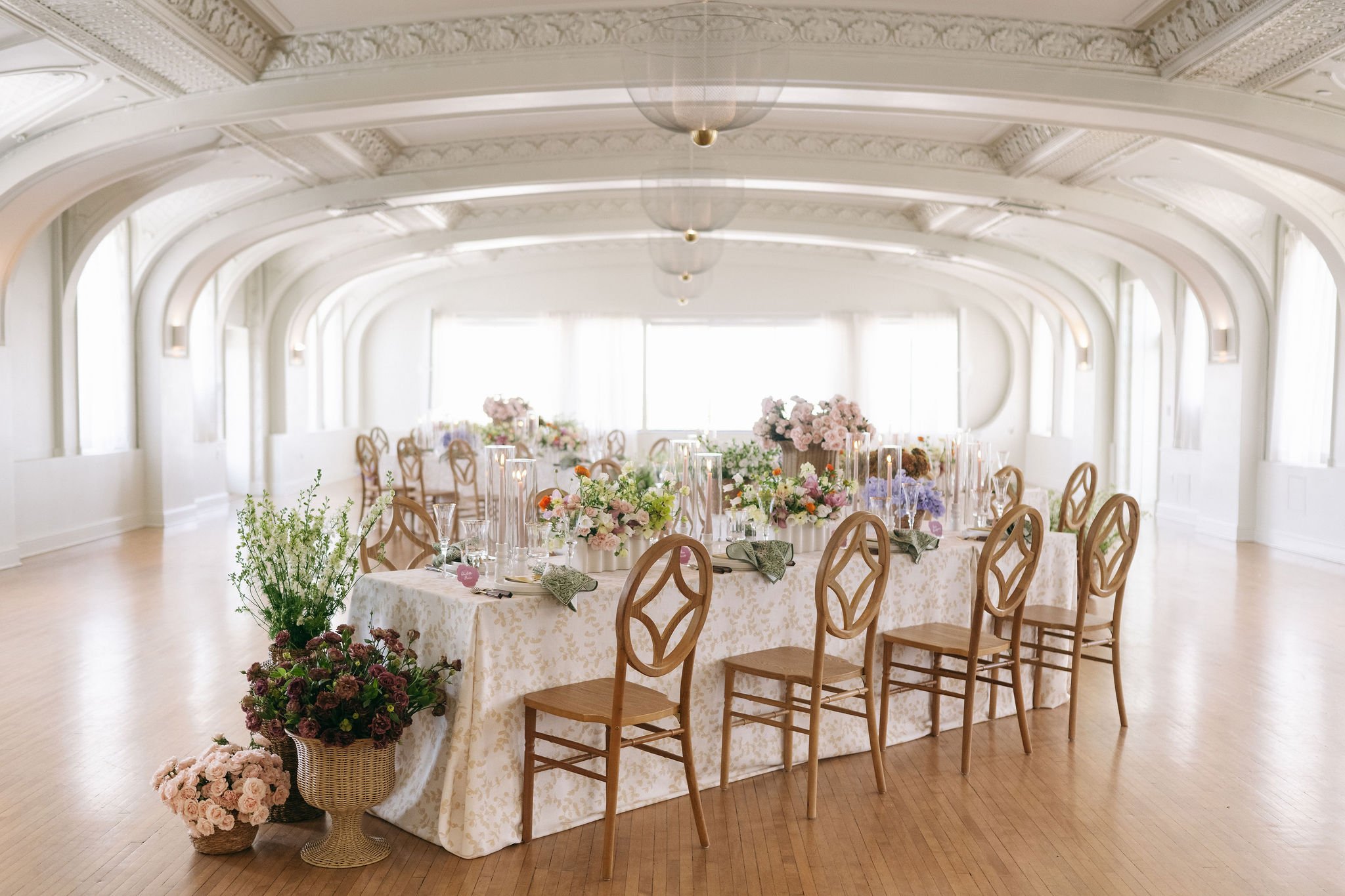 Elegant banquet table decorated with pink, white, and purple flowers in a spacious, bright room with arched windows and ornate ceiling moldings.
