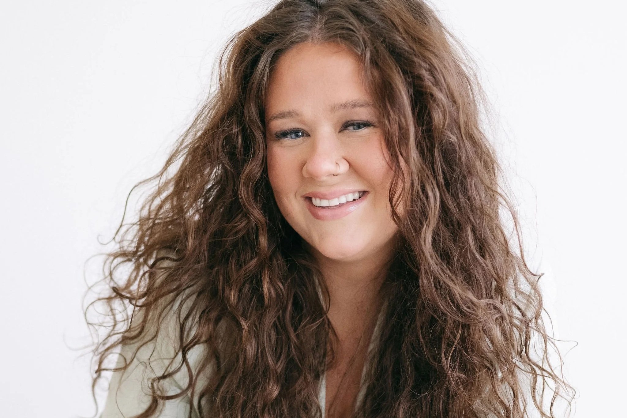 A woman with long, curly brown hair smiling and looking at the camera against a plain white background.