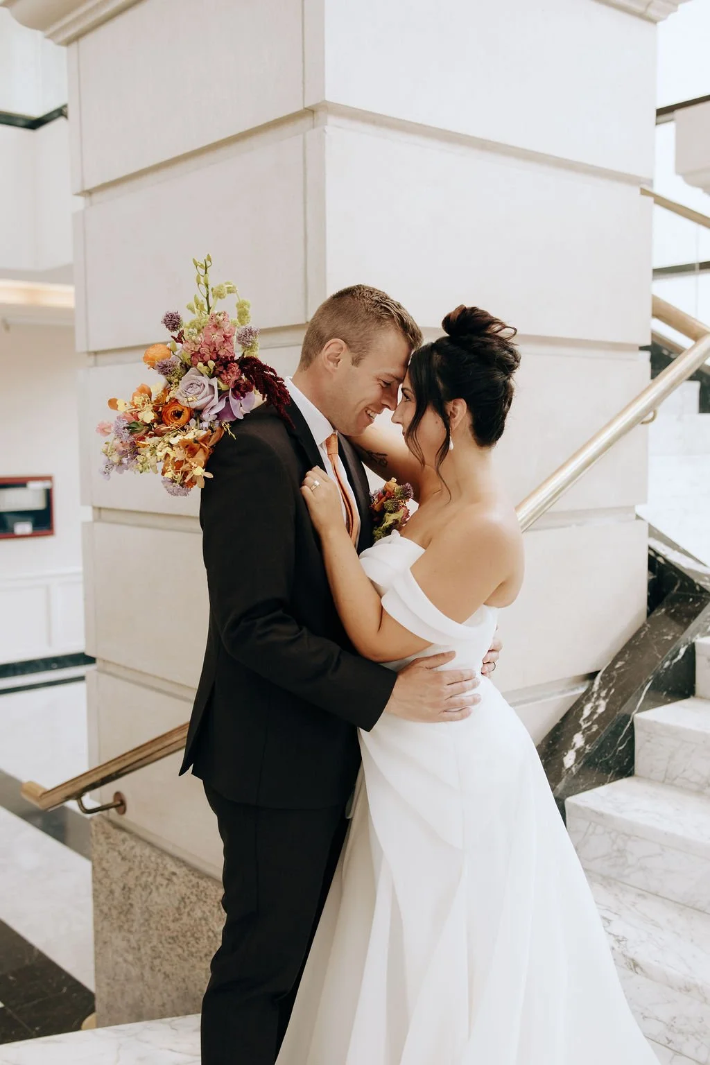 A bride and groom embrace each other in a happy moment on their wedding day indoors. The bride wears a white off-the-shoulder wedding gown and the groom wears a black suit. The bride holds a colorful bouquet of flowers over her shoulder, and they are
