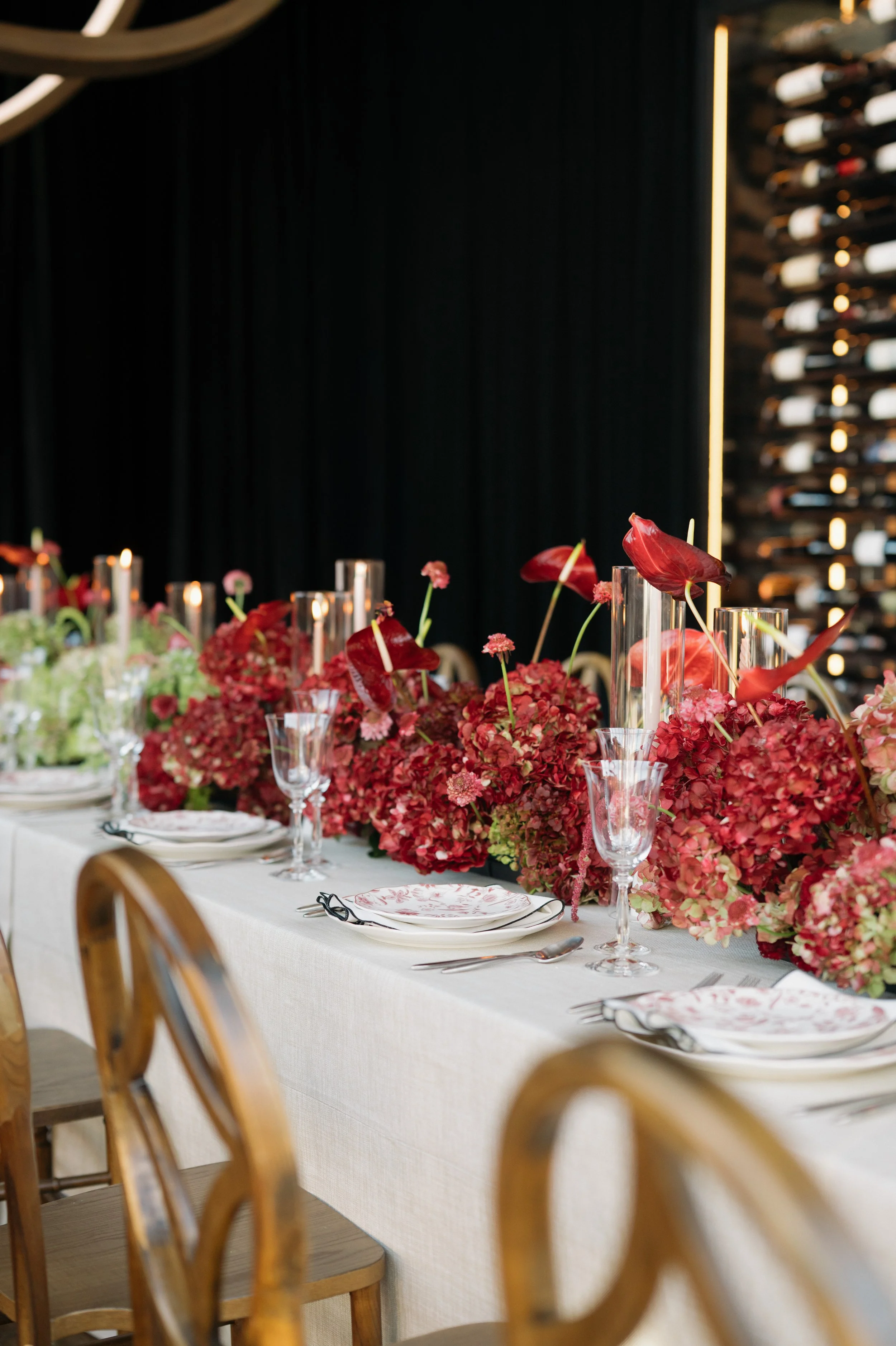 A formal dining table decorated with pink and red floral centerpieces, glassware, and plates for a celebration, with a wine wall in the background.