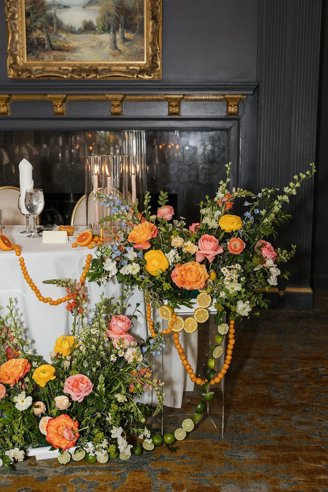 Colorful floral arrangement with pink, yellow, and orange flowers interspersed with greenery, decorated with orange beads and slices of lemon and lime, placed on the floor beside a table set for a formal event.