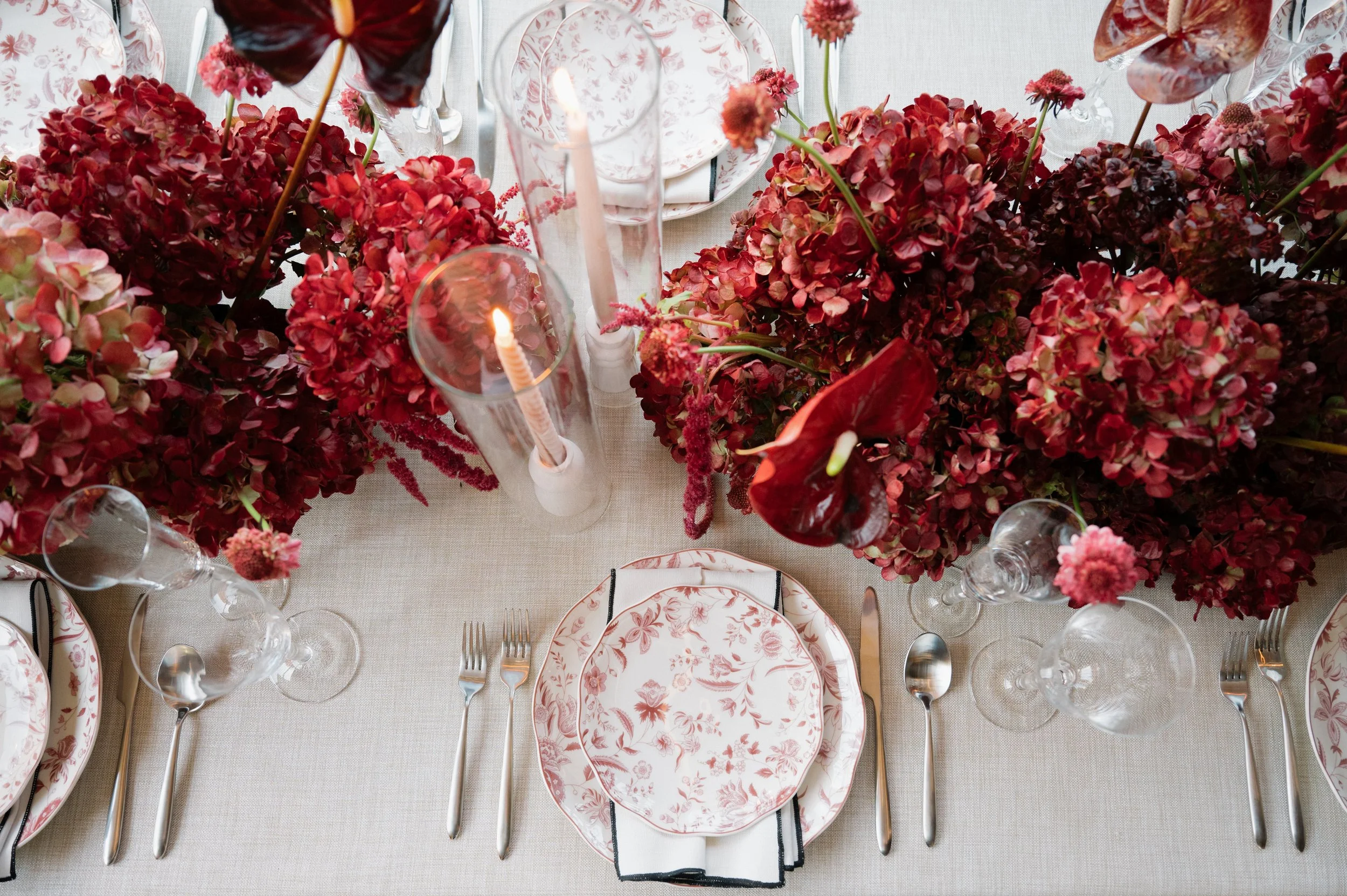Elegant dining table with a flower centerpiece of red and pink hydrangeas and calla lilies, set with pink patterned plates, silverware, and wine glasses, lit candles in glass holders, on a light-colored tablecloth.
