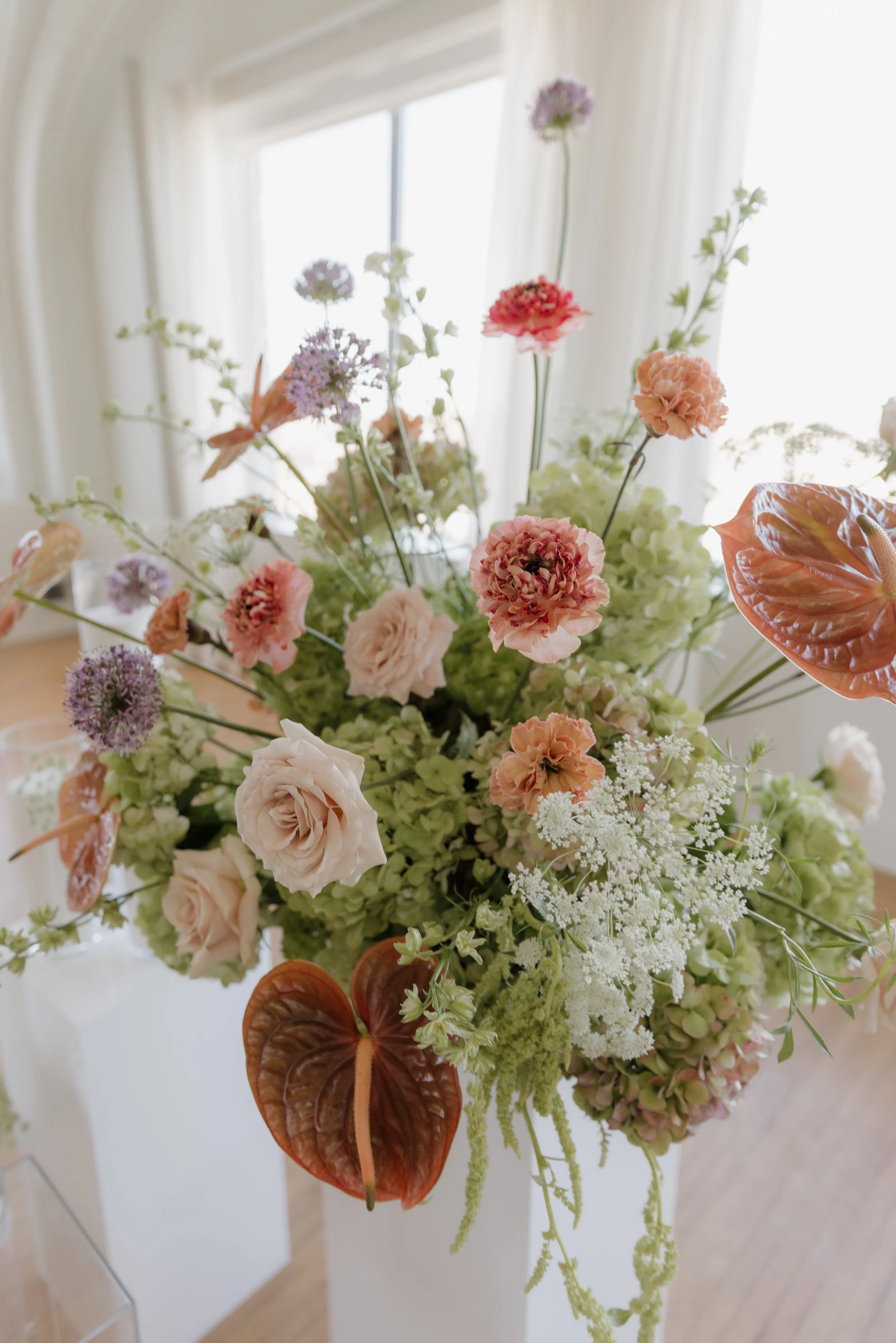 A colorful arrangement of various flowers, including roses, hydrangeas, and tropical foliage, in a white vase near a window.