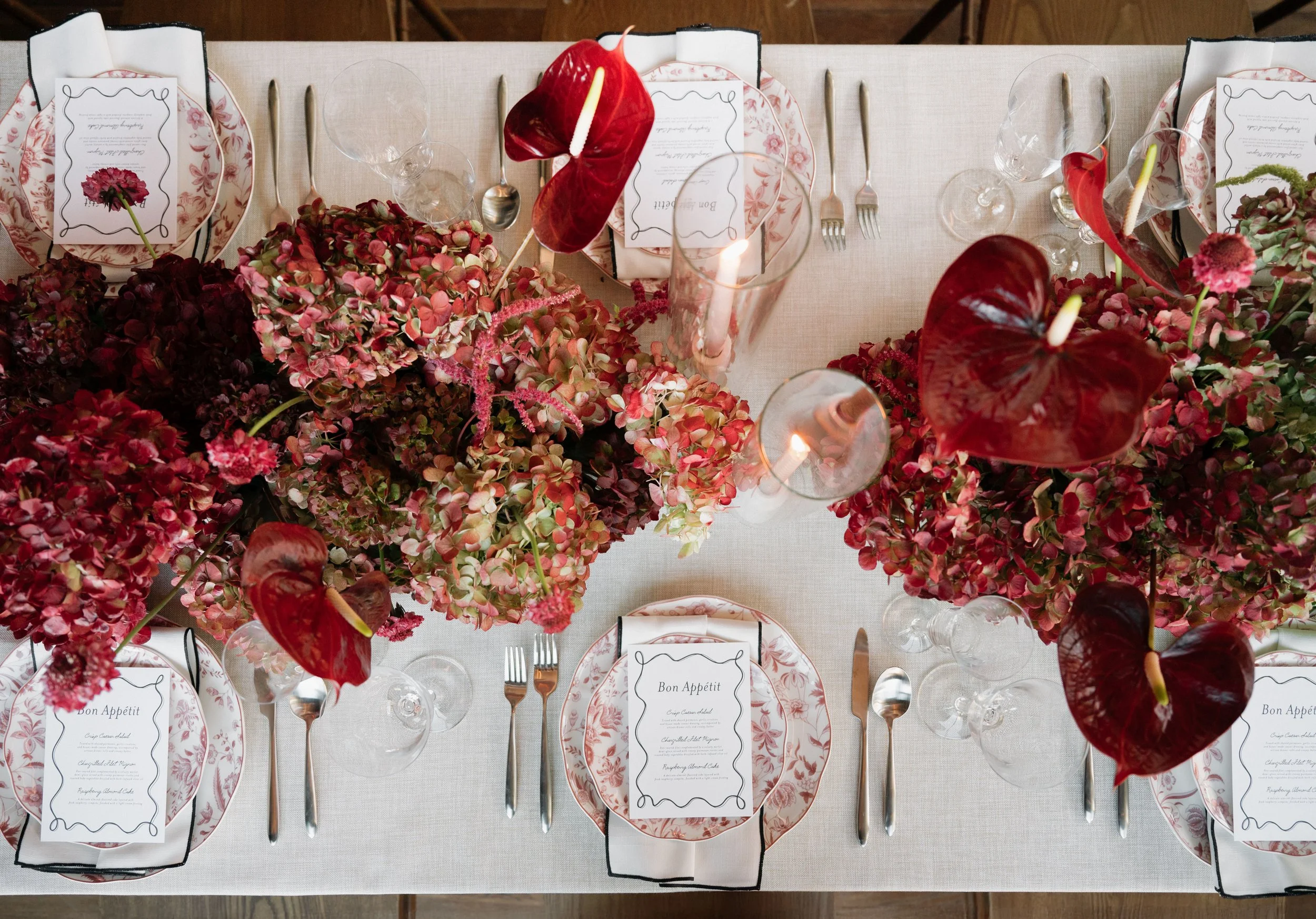 A formal dining table decorated with a floral centerpiece consisting of red and pink hydrangeas, anthuriums, and other flowers. The table is set with plates, silverware, wine glasses, and printed menus at each place setting. There are also lit candles in glass holders.