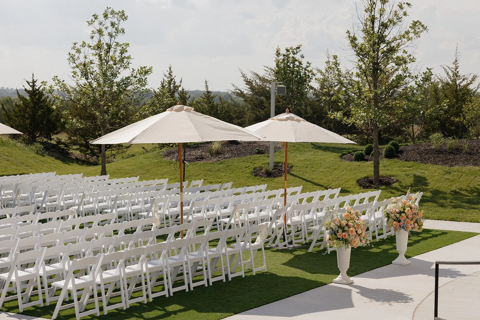 Outdoor wedding setup with rows of white chairs, large white umbrellas, and floral arrangements in tall white vases, surrounded by greenery and trees.