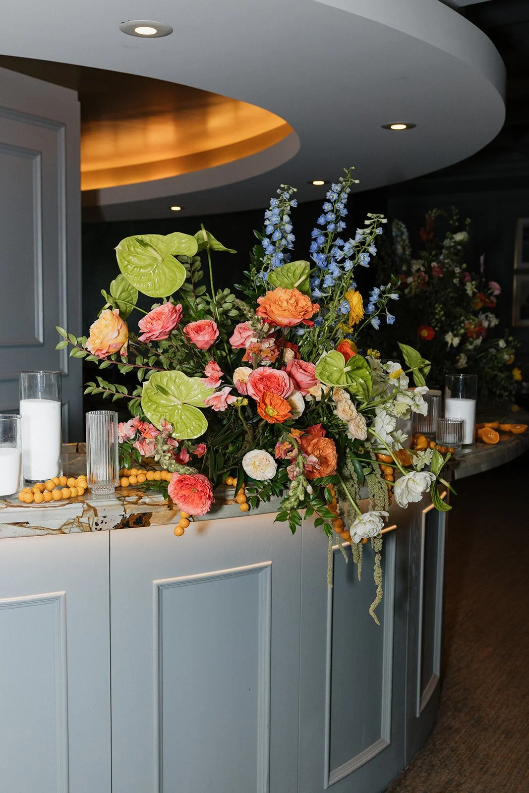 A colorful floral arrangement on a curved marble countertop with candles in glass holders and orange beads.