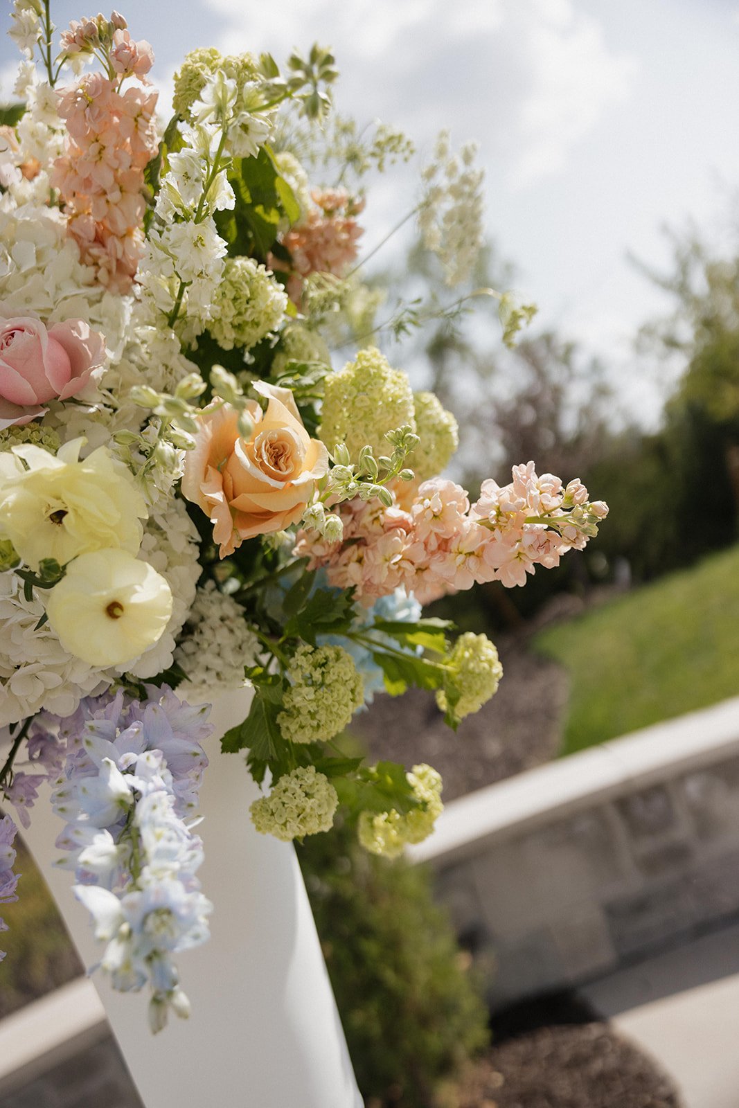 Close-up of a colorful flower arrangement with roses, delphiniums, and other flowers in a white vase outdoors on a sunny day