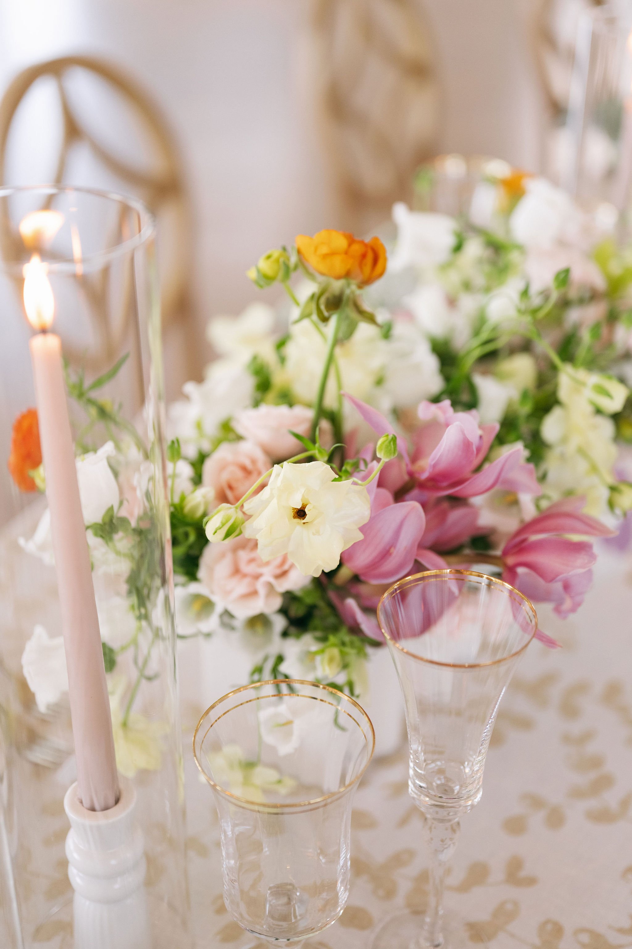 A floral centerpiece with pink, white, and yellow flowers on a table featuring glassware with gold rims and a tall pink candle in a glass holder, set on a cream-colored tablecloth with a subtle pattern.