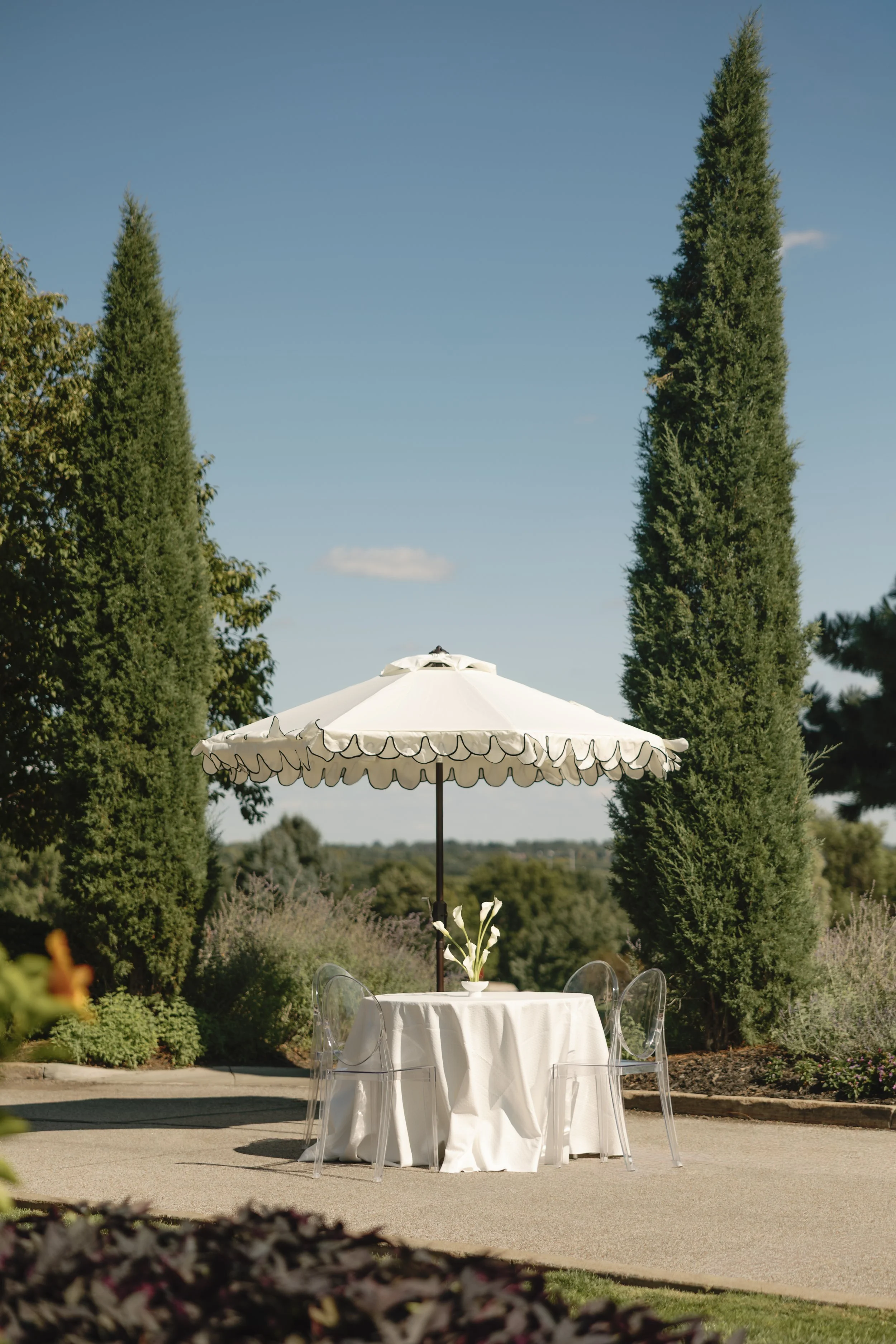 An outdoor table with a white tablecloth and a white umbrella, surrounded by transparent chairs, set in a garden with tall green trees under a blue sky.