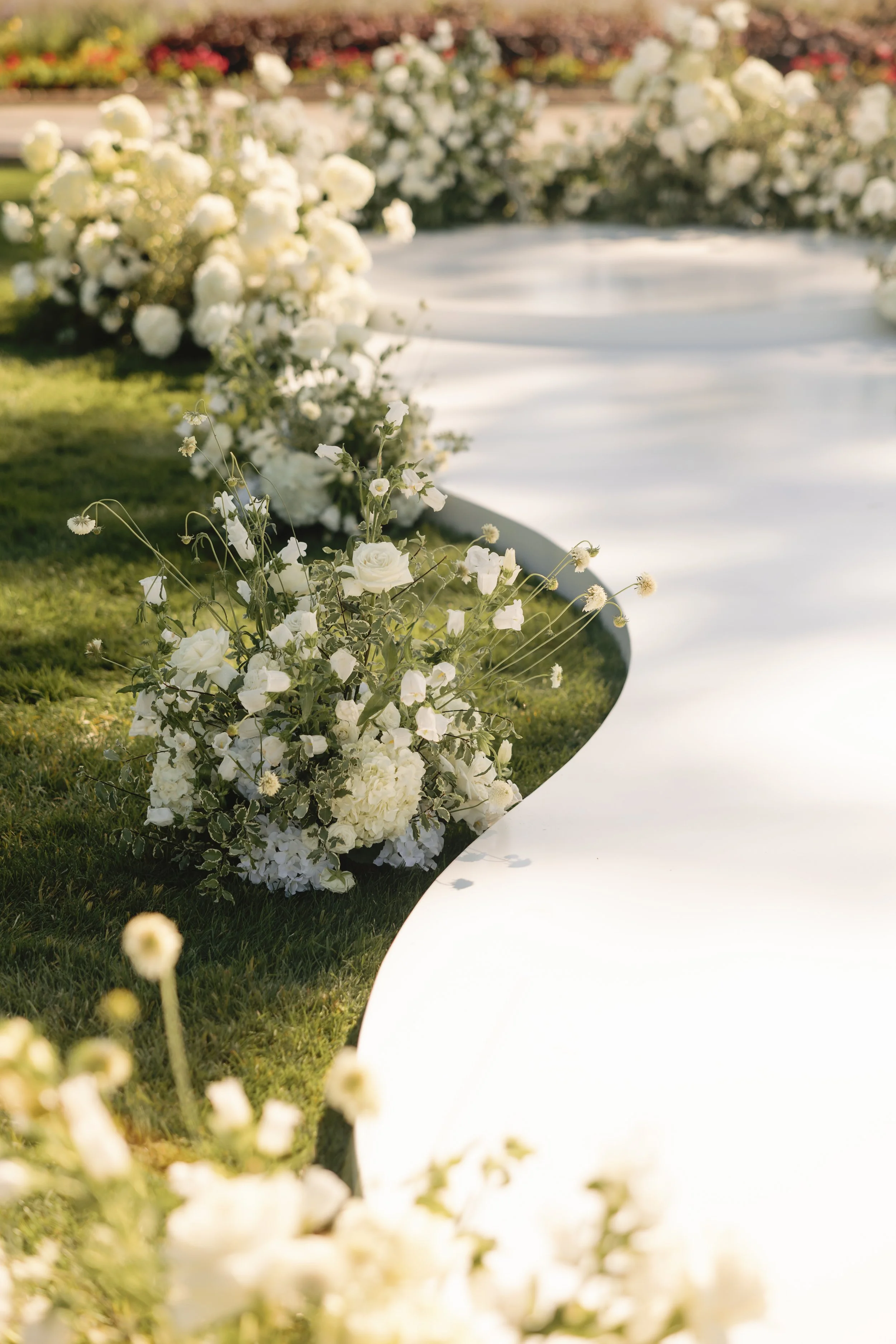 White floral arrangements and greenery along a curved white pathway in an outdoor wedding setting.