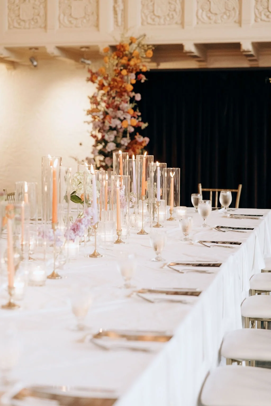 Elegant banquet table setup with candles, glasses, and floral decorations in front of a black curtain and a floral backdrop.