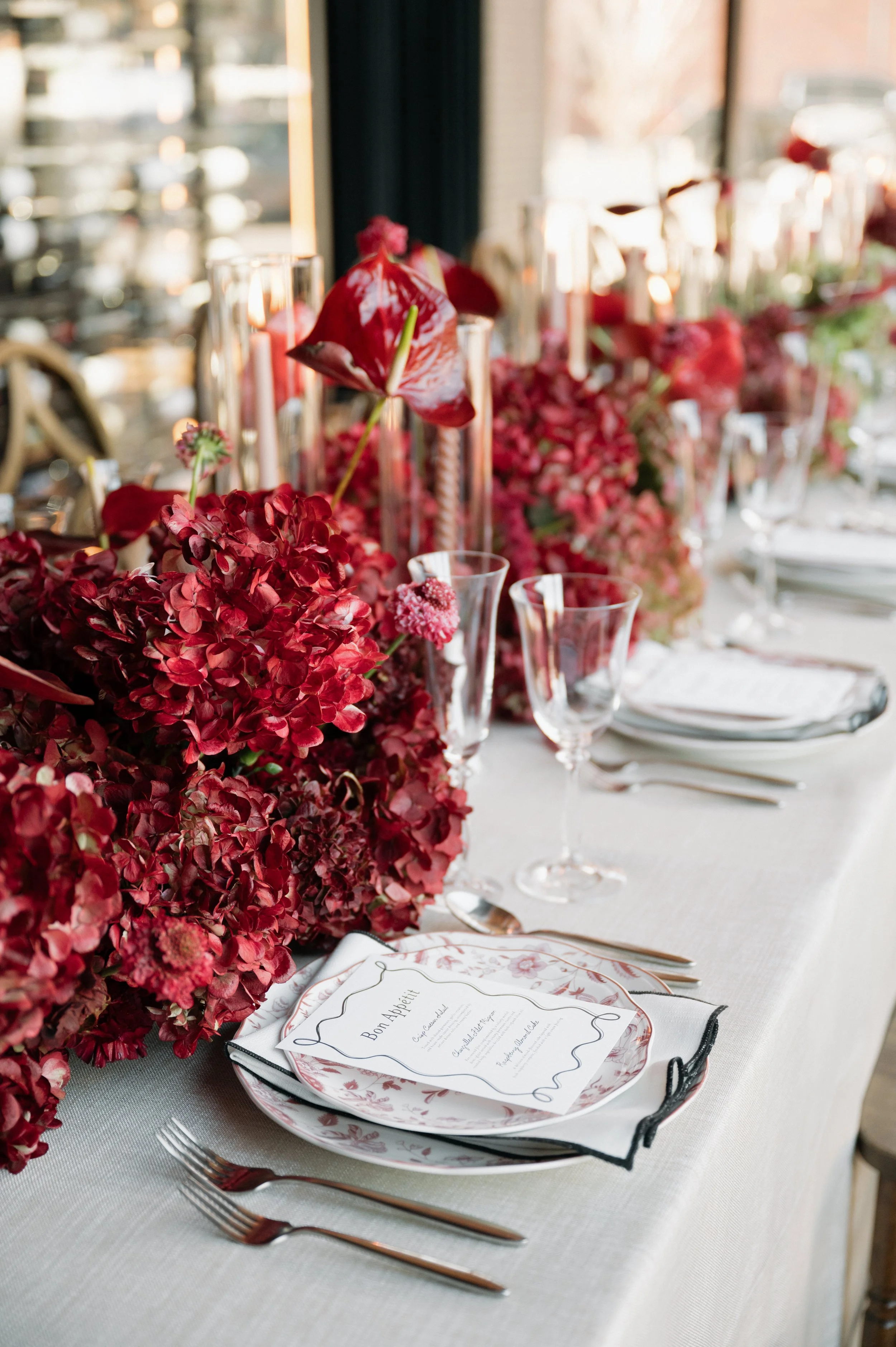 Elegant wedding table decor with a long floral centerpiece of red and pink flowers, tall glass candle holders with red candles, a place setting with a decorative plate, a printed menu, and silverware on a white tablecloth.