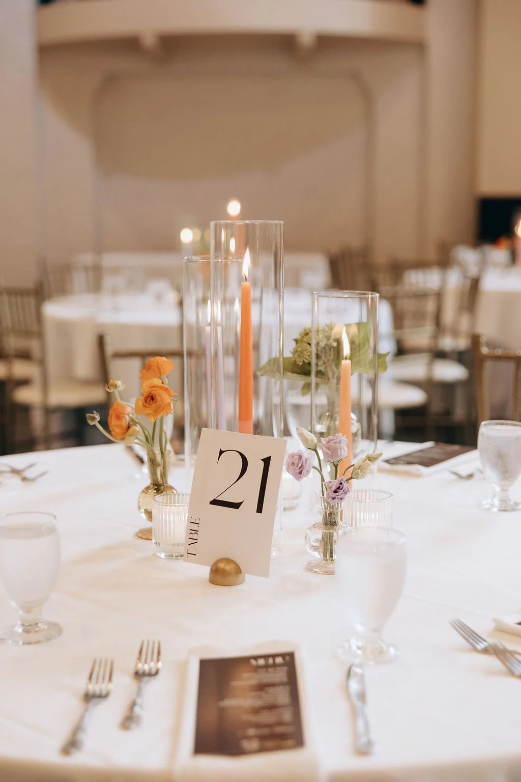 A wedding table setting with a floral centerpiece, tall candles in glass holders, numbered table card '21', and place settings with water glasses and silverware in an elegant venue.