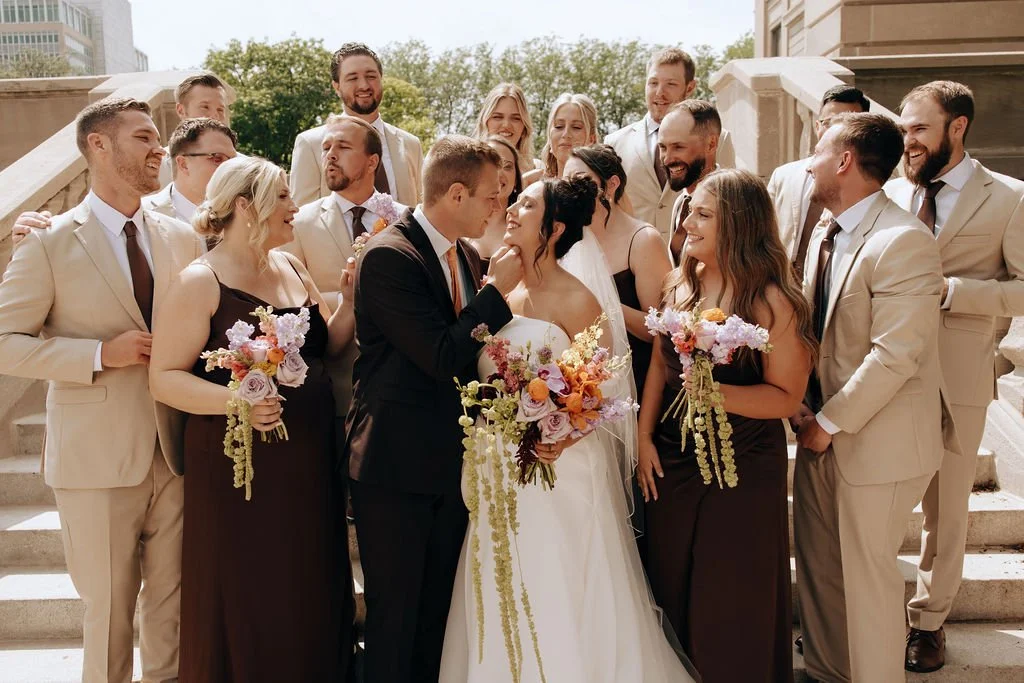 Bride and groom surrounded by bridesmaids and groomsmen on steps outside, smiling during a wedding celebration.