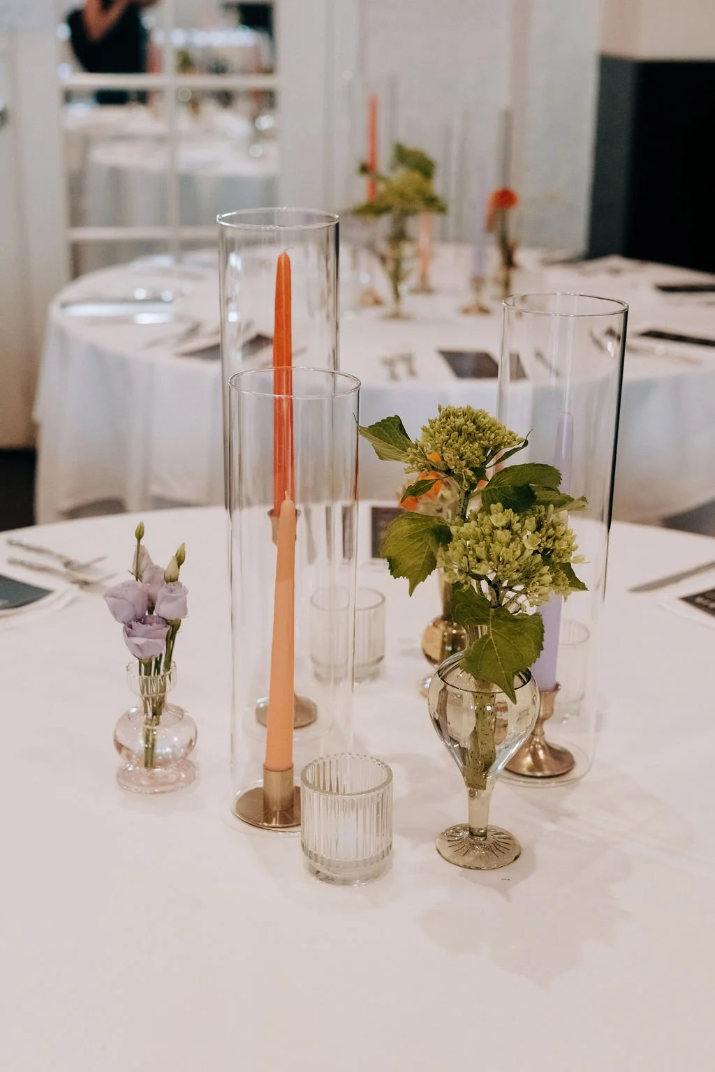 A decorated table set for a formal event or dinner with glass vases holding flowers and candles, arranged on a white tablecloth.