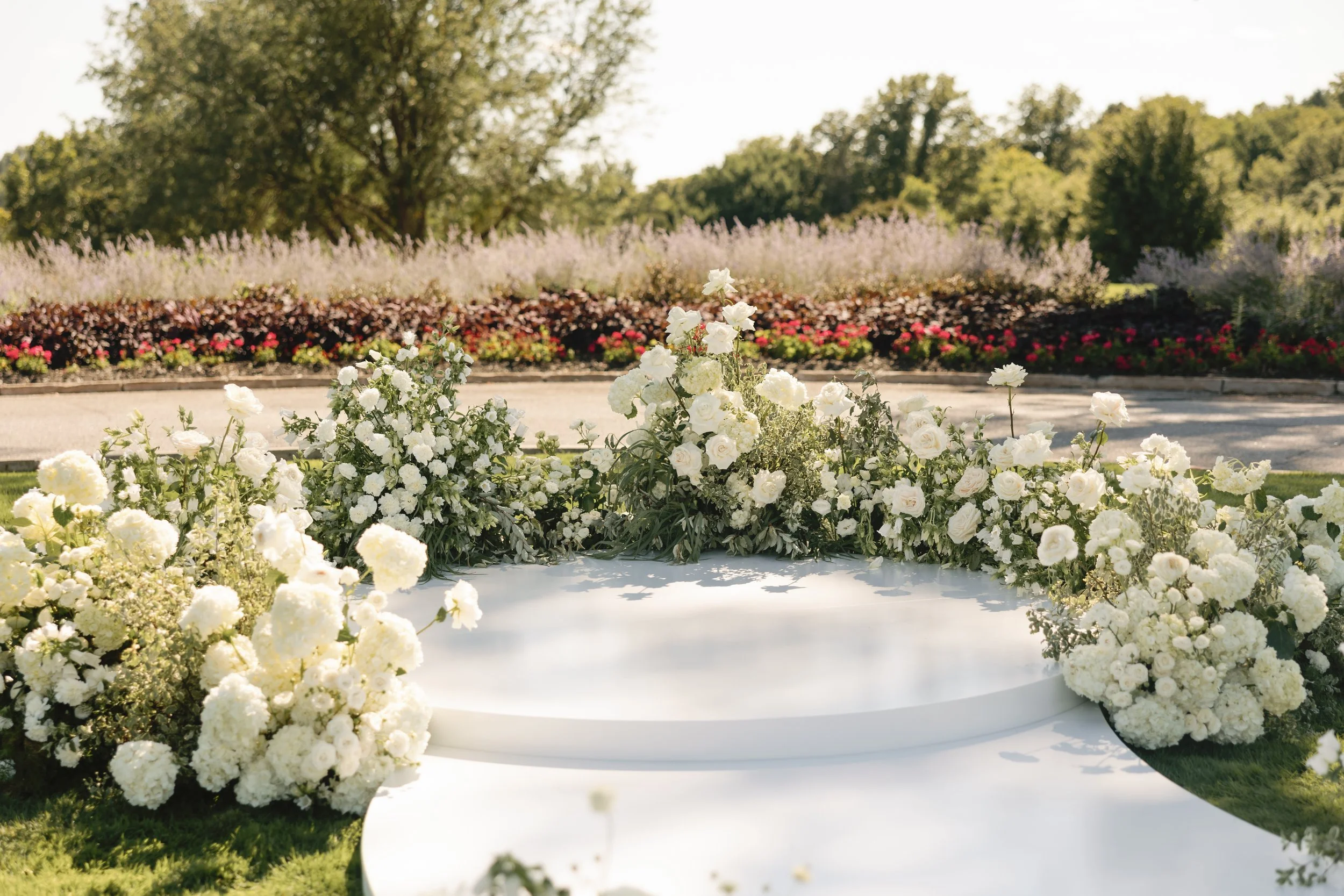Wedding altar decorated with white flowers, set outdoors in a garden with trees and colorful flowers in the background.