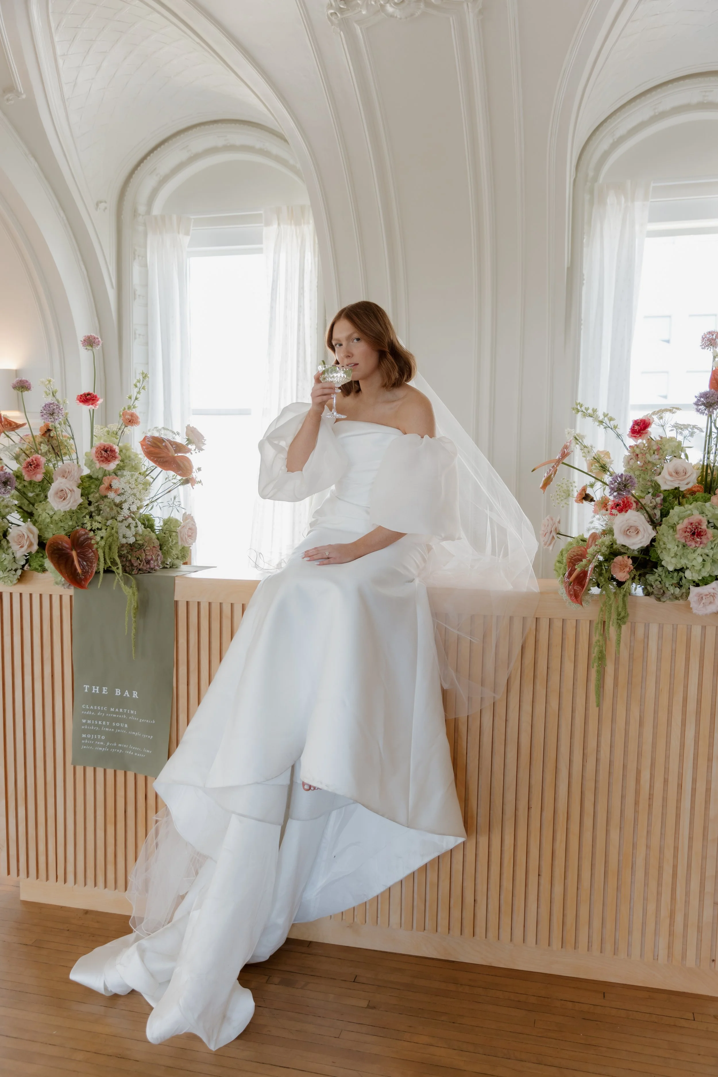 A woman in a white wedding dress sitting on a wooden bar counter, holding a cocktail glass, with floral arrangements on either side in a bright, elegant room with large windows.