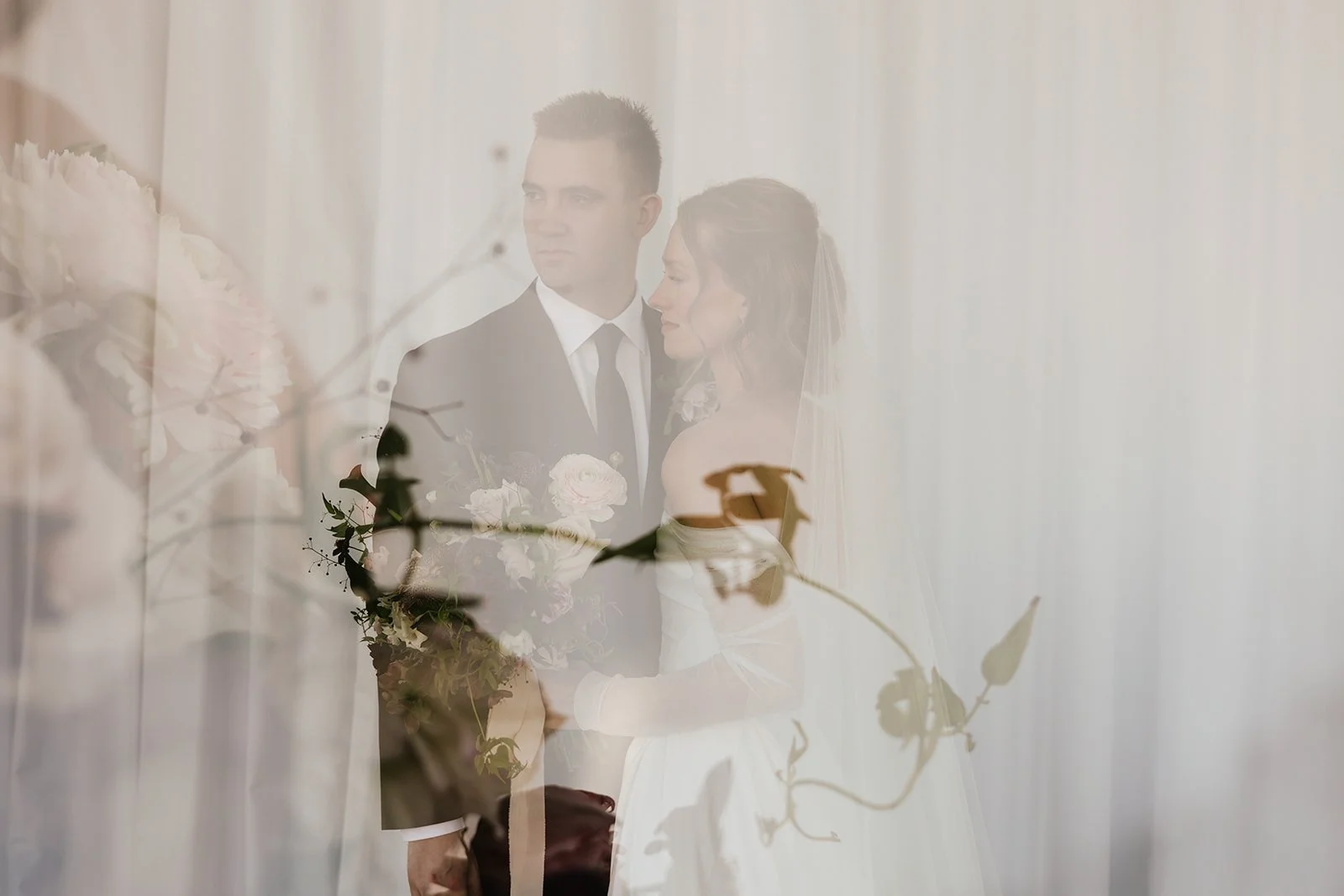 A couple dressed in wedding attire standing close together, with the woman holding a bouquet of flowers, seen through a semi-transparent curtain or screen with floral and leafy patterns.