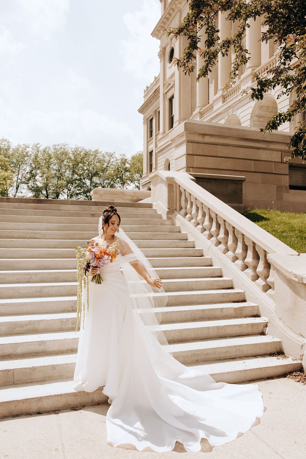 A bride in a white wedding gown with a long train, holding a bouquet of colorful flowers, standing on an outdoor staircase in front of a large, elegant building with columns and decorative architecture, during daytime.