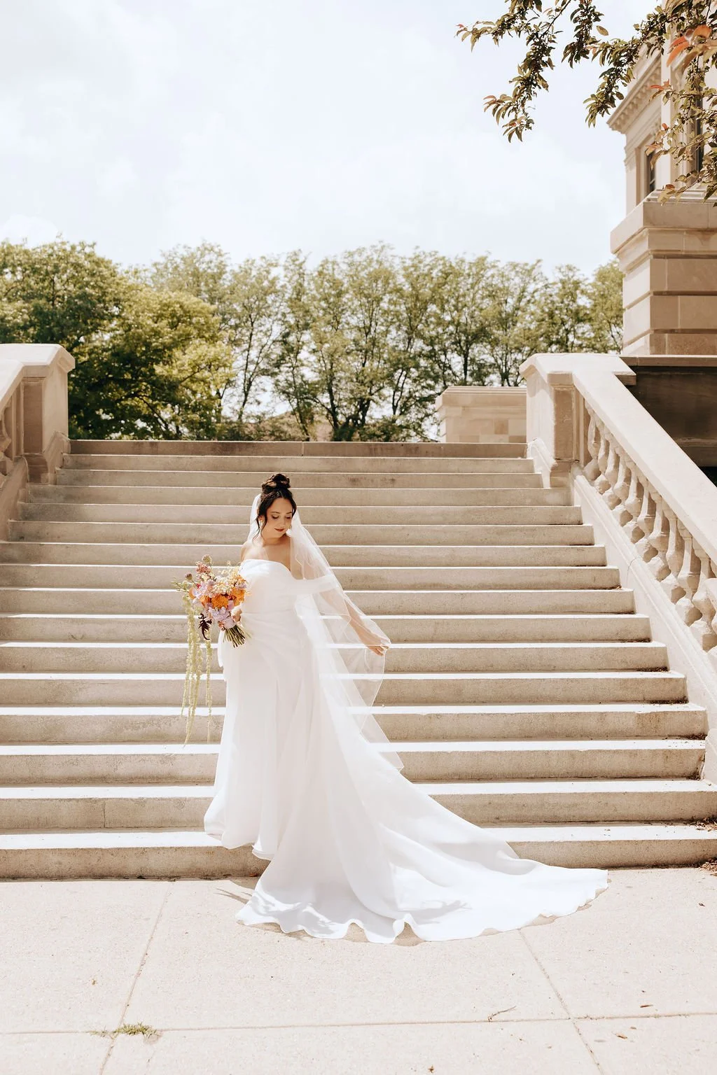 A bride in a white wedding gown holding a bouquet of flowers, standing on outdoor stairs with trees and a building in the background.