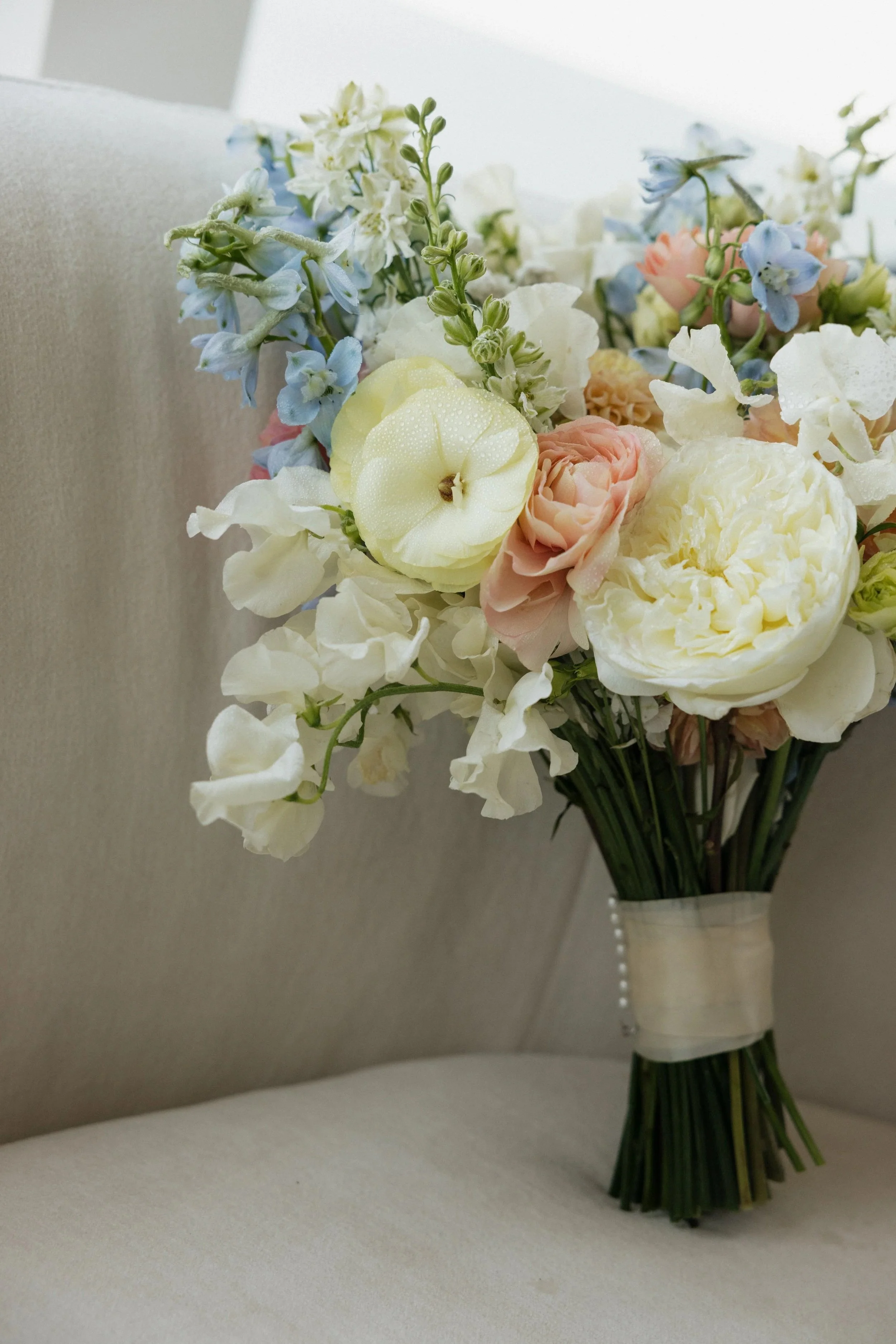 A bouquet of pastel-colored flowers, including white, pink, yellow, and blue blooms, arranged in a cream-colored vase, set on a light-colored surface against a neutral background.