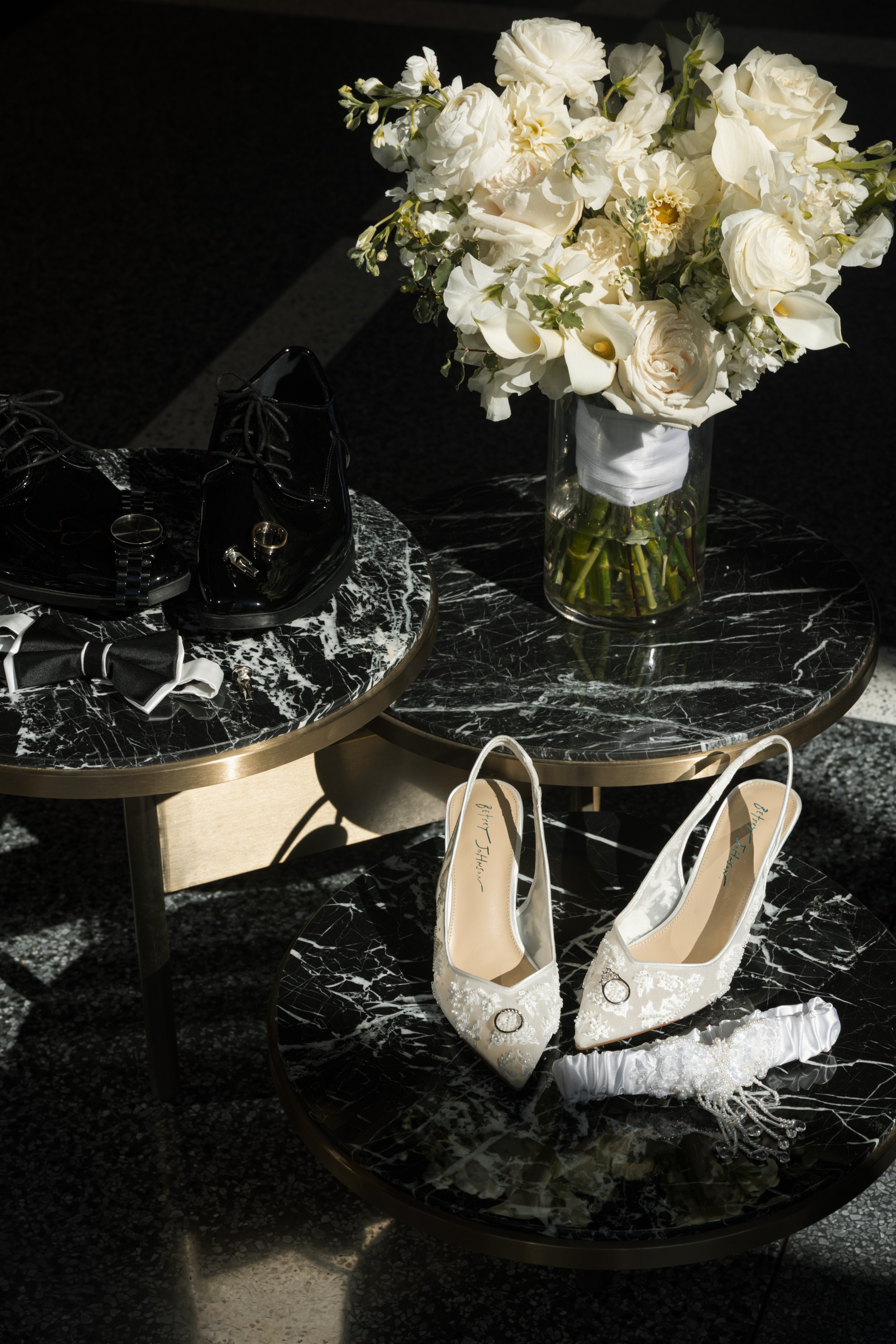 Wedding shoes, bouquet of white flowers, in a glass vase, resting on black marble side tables.